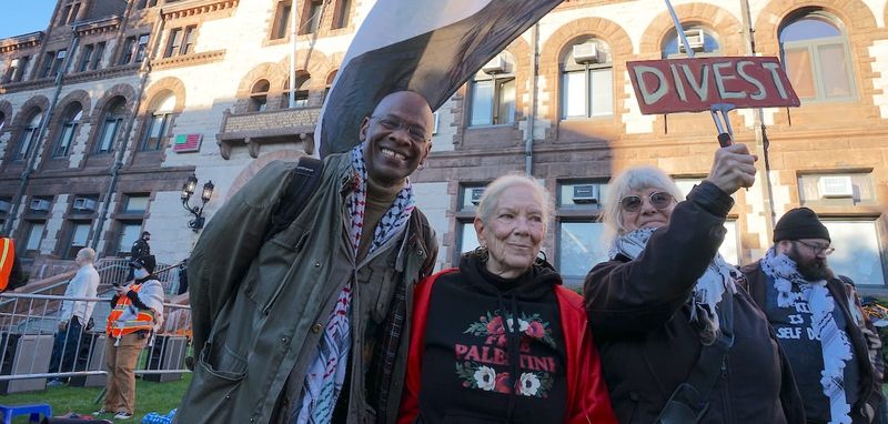 Ann Antonellis holds a sign that reads "divest", attached to a Palestinian flag, standing next to Michel DeGraf and Susan Escovitz in front of Cambridge City Hall.