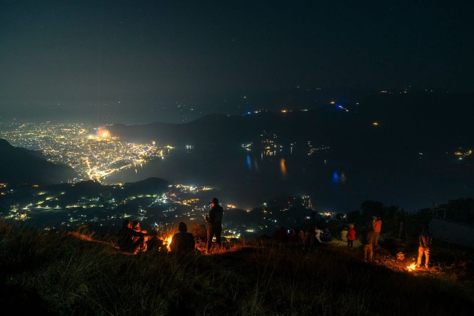 Photo of several people around two campfires on a grassy hill at night, overlooking a bright cityscape in the valley below.