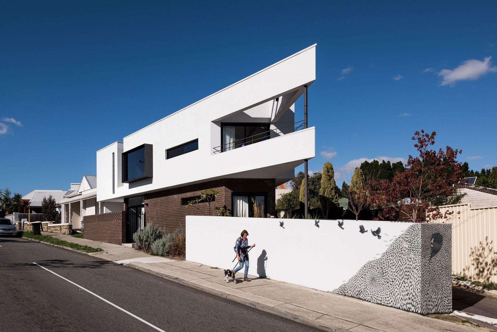 Street facing facade of a triangle-shaped, multistory home. Ground floor constructed of brick and a white, rendered second story. 