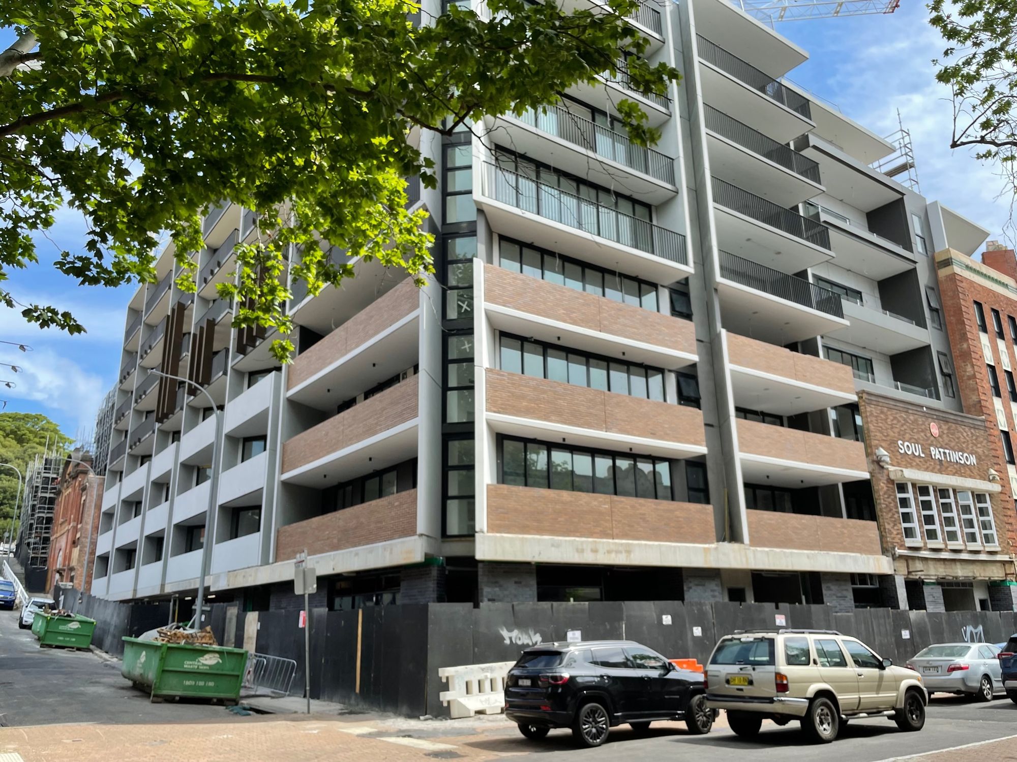 A street-level view of a modern multi-story apartment or office building. The structure features a contemporary design with a mix of gray and wooden facade elements, interspersed with generous balconies on multiple levels. Large glass windows punctuate the building, allowing ample natural light. Adjacent to this new structure is an older brick building with a sign that reads "SOUL PATTINSON". A lush green tree canopy at the top left adds a touch of nature to the urban scene. In the foreground, there's ongoing construction or renovation evident from the green waste bins, wooden barricades, and construction-related materials. Parked along the road are several vehicles, indicating a busy city environment. The image contrasts the evolution of architectural styles and the blend of the old with the new in a bustling urban setting.
