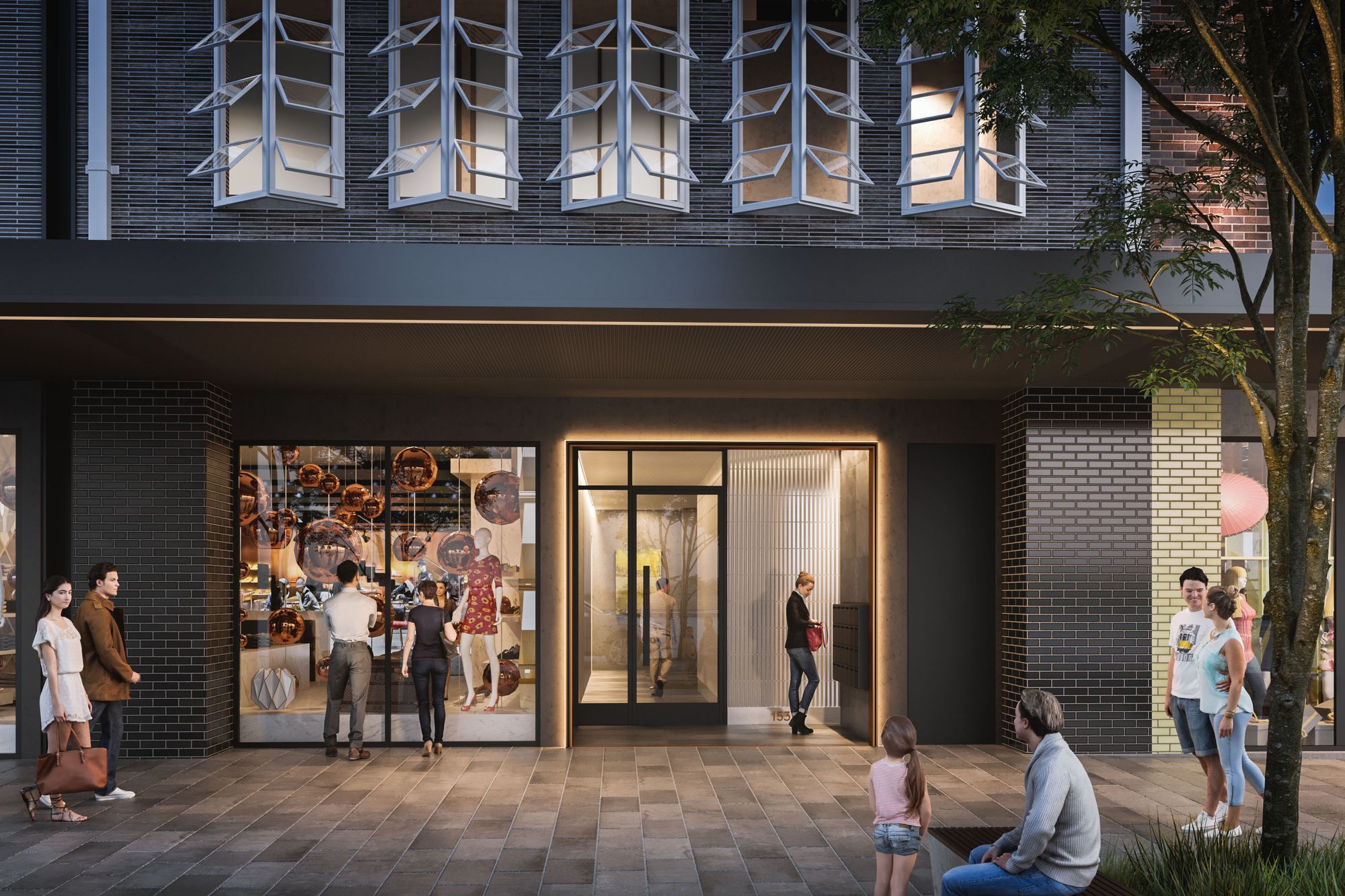 The image depicts an urban street scene in front of a contemporary building. The building's façade showcases a mix of brickwork and geometric window designs. On the ground level, there is a shop with a transparent glass front that displays various stylish items, possibly related to fashion or decor. Several pedestrians, including couples and individuals, are walking by or standing nearby, interacting or observing the shop's offerings. A tree stands adjacent to the structure, and there's a small seating area where a man and a young child are seated. The overall mood appears to be calm and relaxed, capturing a typical evening in a modern city setting.