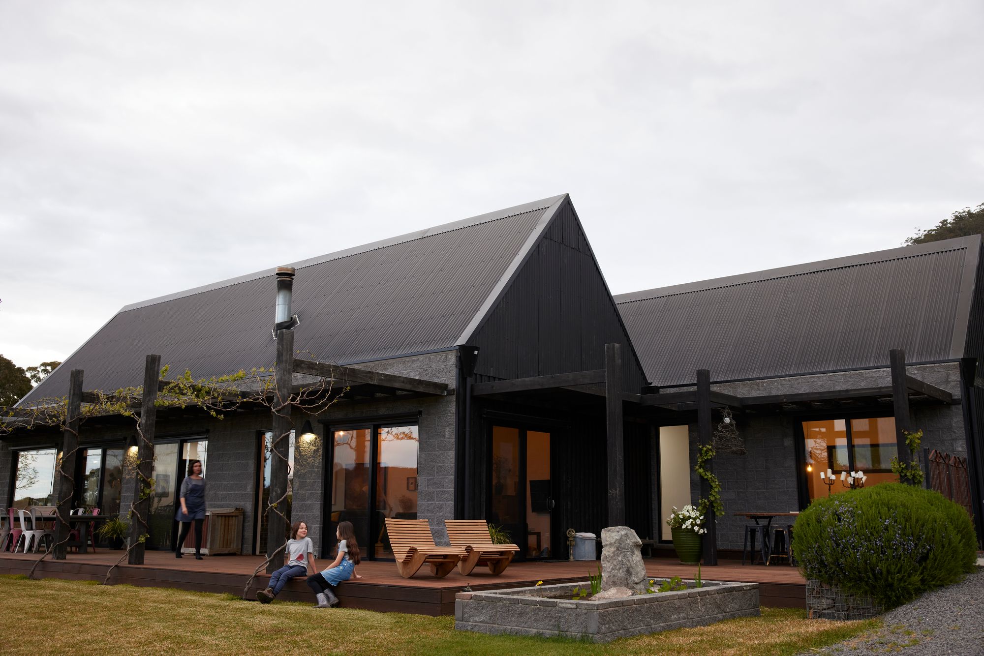 The building's façade is composed of dark materials, possibly wood or a wood-like finish, which contrasts sharply with the light trim around the windows and doors. It has a rustic charm with exposed beams supporting an overhang that shelters an outdoor seating area. The landscaping includes manicured bushes and a vine-covered pergola, adding a touch of greenery against the building's dark tones. People can be seen enjoying the outdoor space, indicating that this building may be a public venue like a restaurant or a winery. The sky is overcast, suggesting a cool or cloudy day.