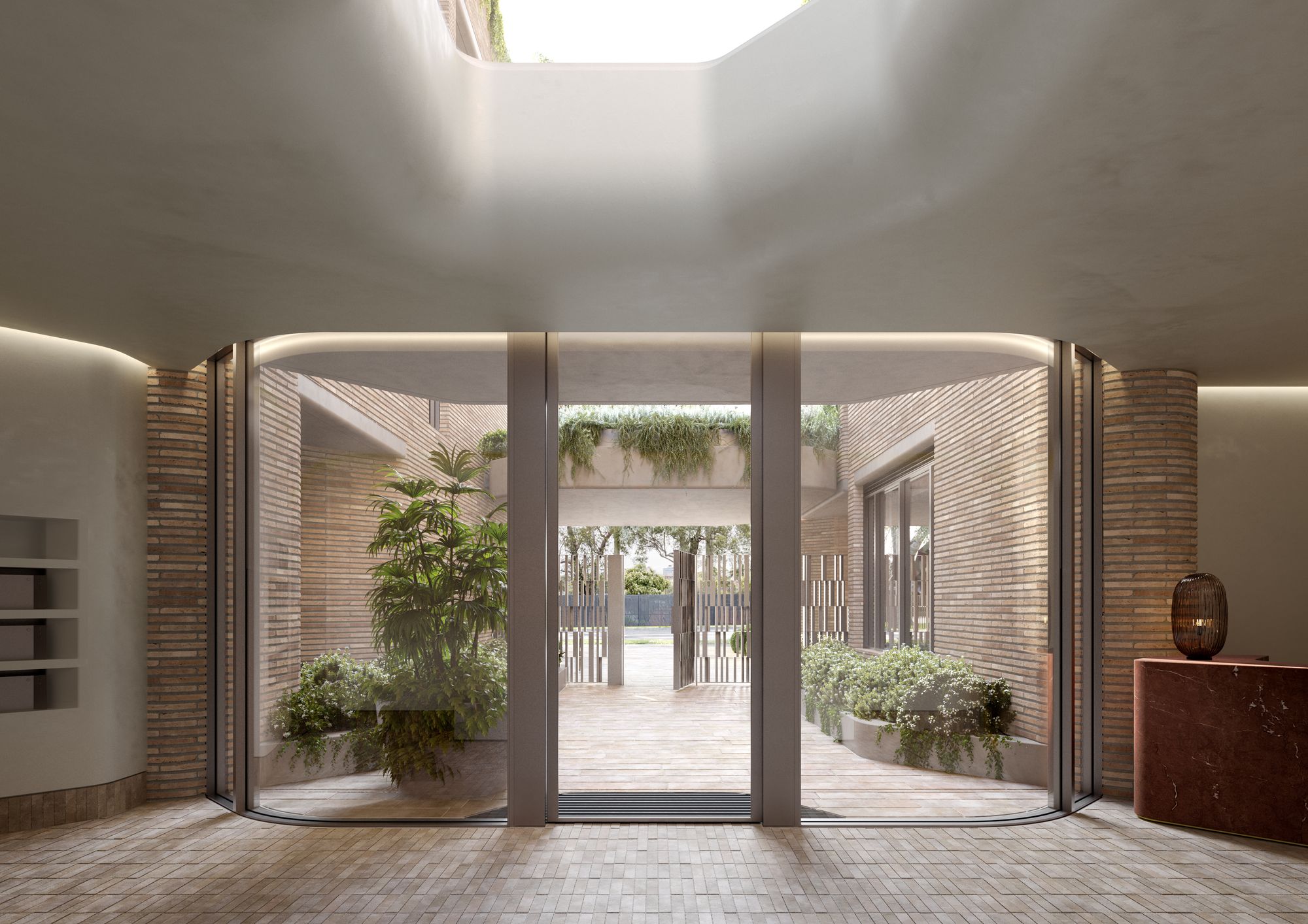 Como Toorak by Prime Edition x Jolson.  This image shows a modern architectural space that blends indoor and outdoor elements. The curvilinear ceiling design creates a smooth transition between the interior and exterior areas. Floor-to-ceiling glass doors enhance natural light and allow a clear view of the outdoor courtyard, which features lush greenery and planter beds, contributing to an inviting and serene atmosphere. The use of bricks on both the exterior and interior walls adds texture and a sense of continuity to the design, while the tiled floor provides a durable and practical surface that complements the earthy tones. 