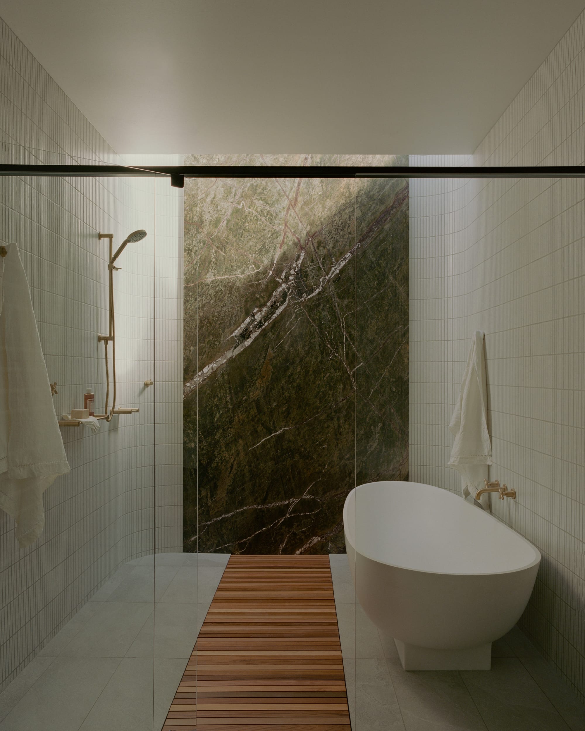 An architectural shot showing a modern white-tiled bathroom with sleek golden fixtures, a large mirror, and a transparent glass shower enclosure revealing a natural rocky landscape outside.