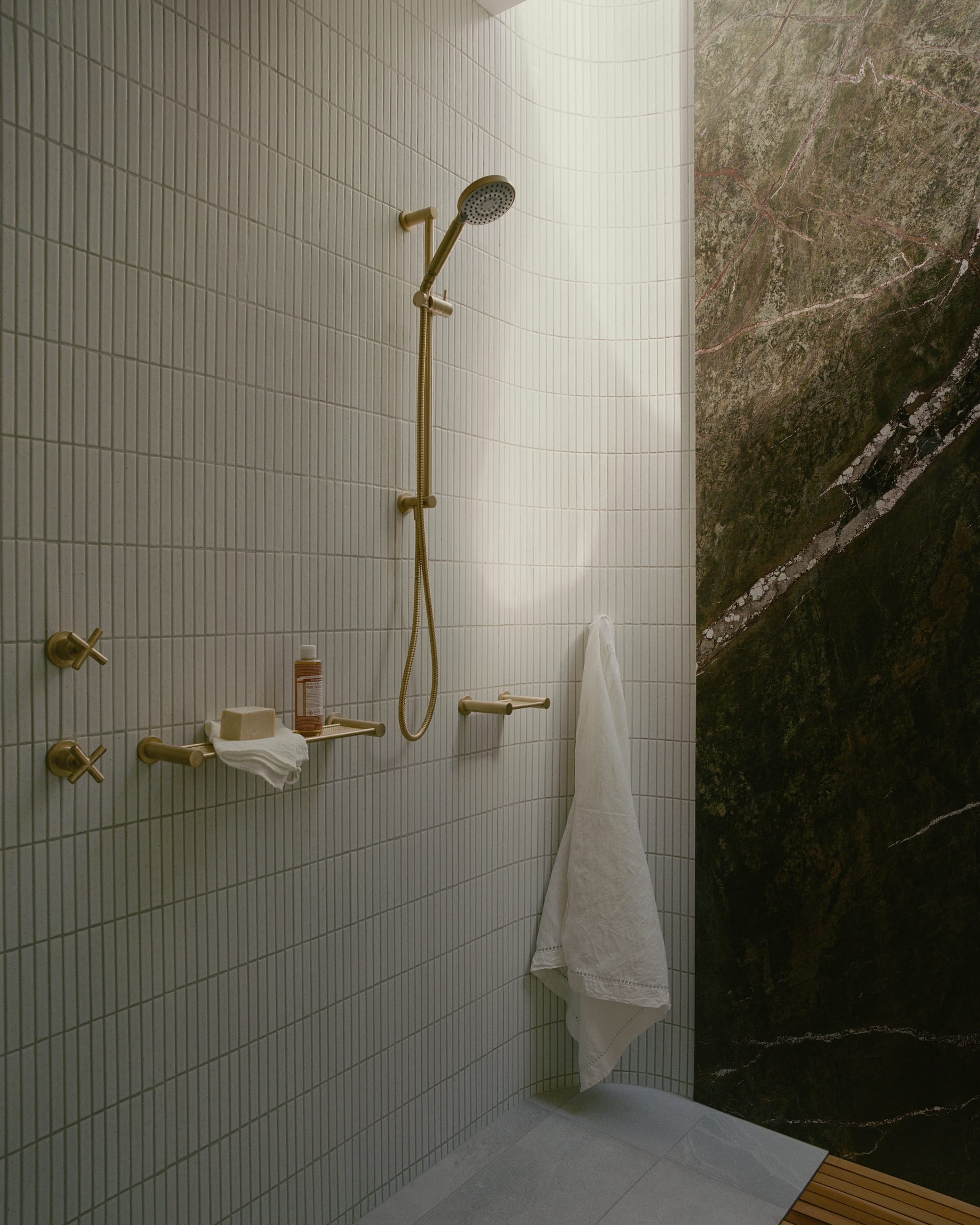 white-tiled bathroom with sleek golden fixtures, a large mirror, and a transparent glass shower enclosure revealing a natural rocky landscape outside.
