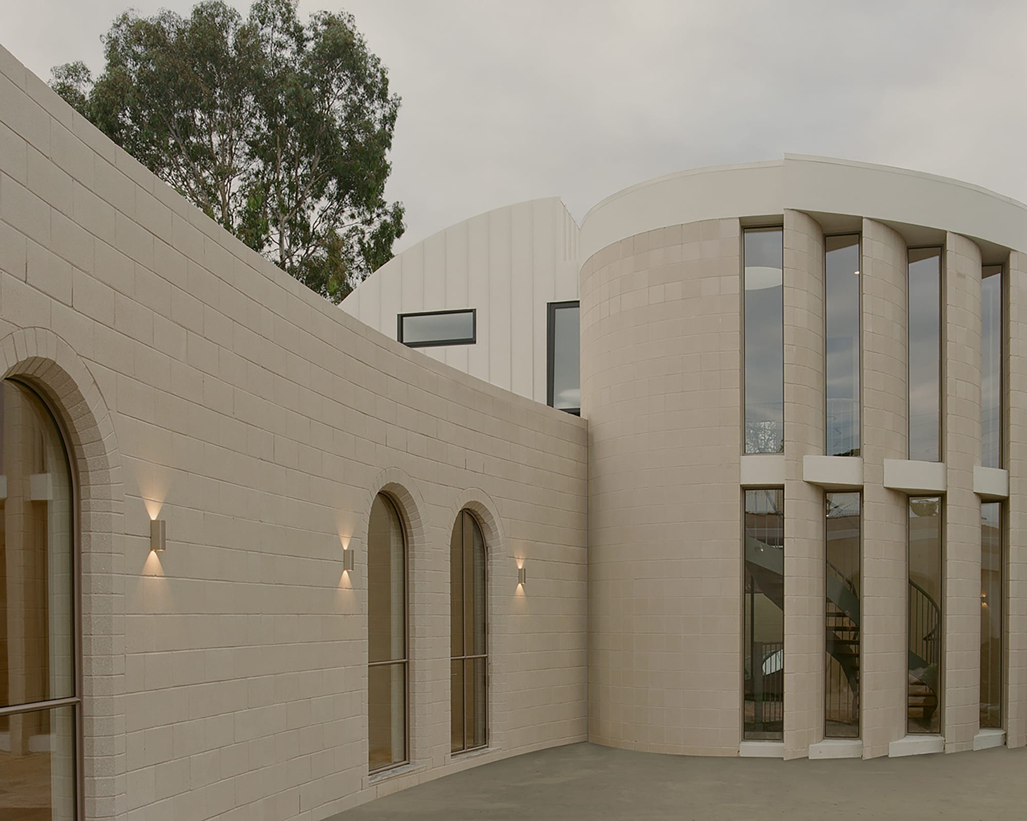 architectural photograph of a modern building featuring curved and straight white walls under an overcast sky. The building has large, vertical windows and wall-mounted lantern-style lights.