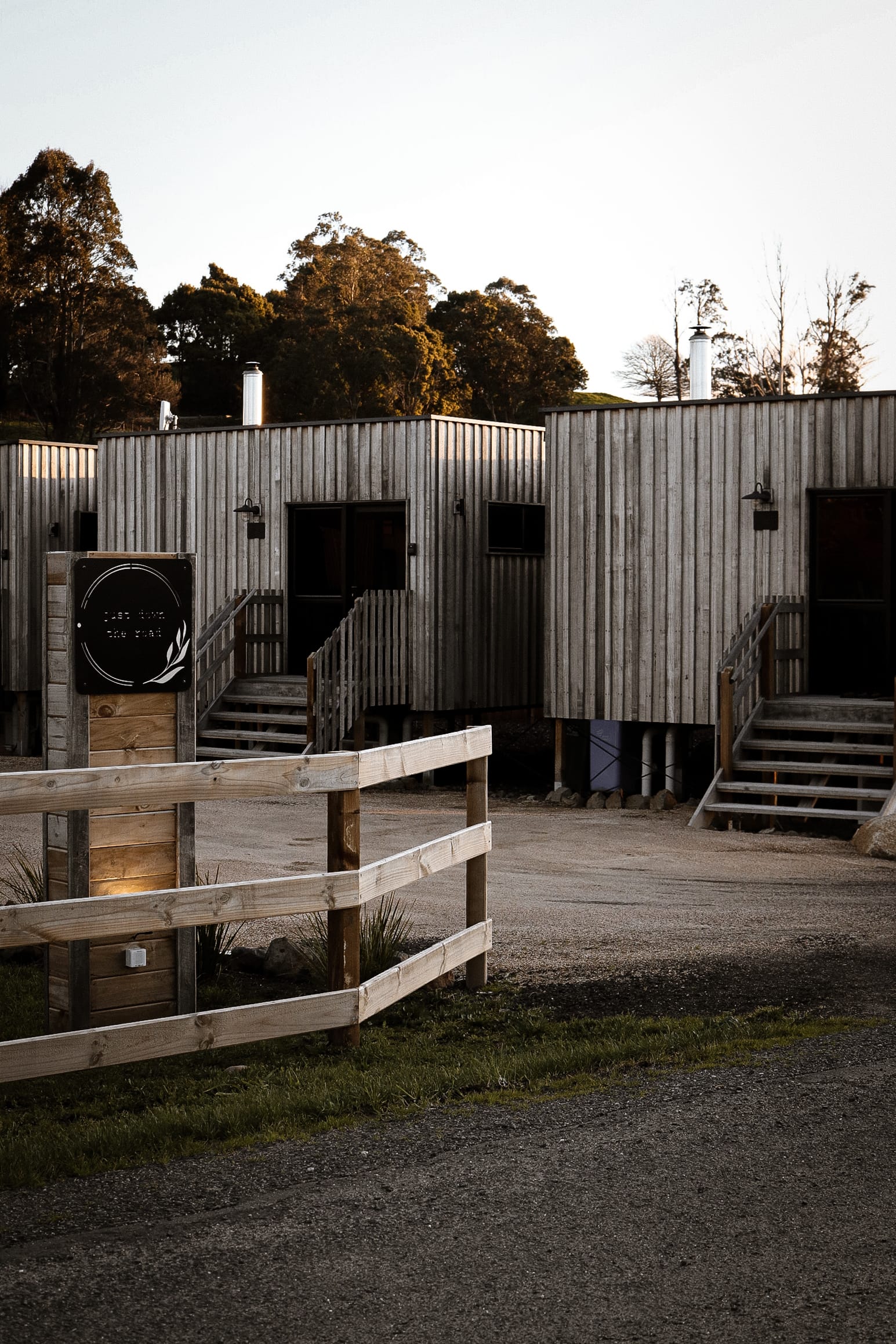 Just Down the Road. Photographer: Studio Winslow.Close-up of the exterior wooden staircase and railing of a modern building with corrugated metal siding, with a gravel ground below and subdued evening light softening the scene.