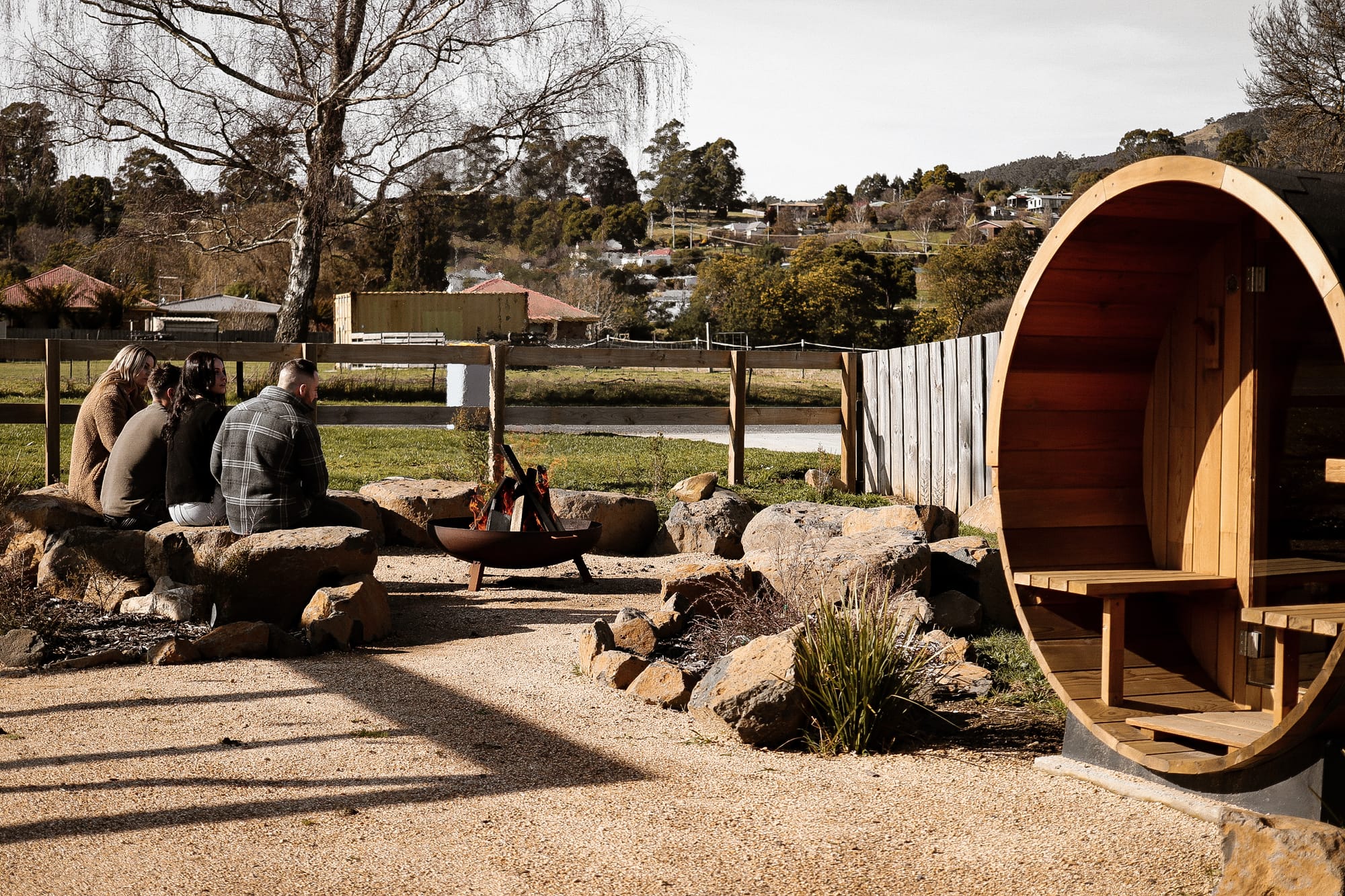 Just Down the Road. Photographer: Studio WinslowThree people sitting by a rock fire pit near a river, with the cylindrical wooden sauna in the background, surrounded by a grassy area and trees under a soft blue sky.