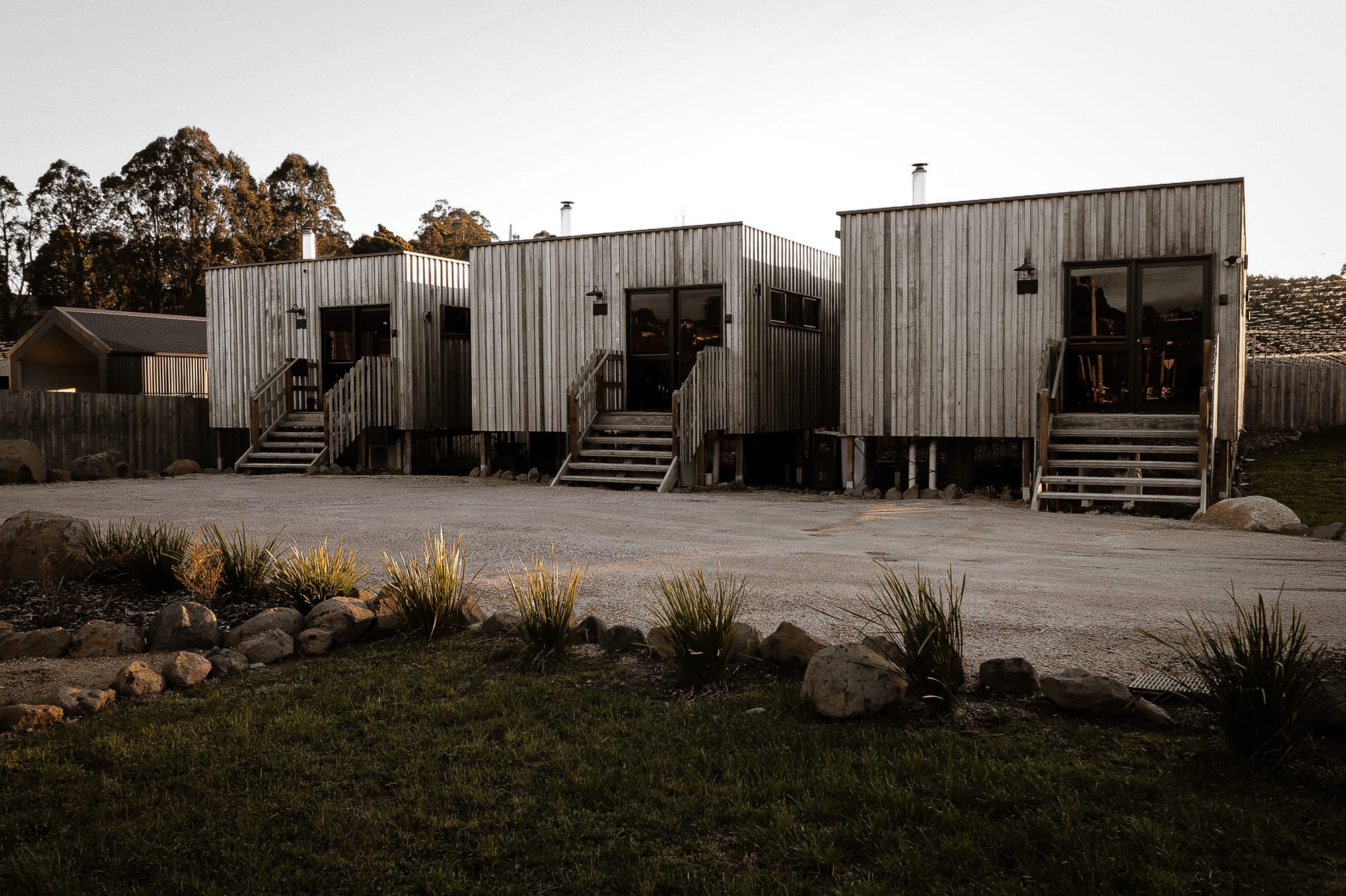 Just Down the Road. Photographer: Studio Winslow. A twilight view of modern rural accommodation featuring two-story buildings with corrugated metal siding and wooden staircases, set in a landscape with a gravel driveway and a garden with small shrubs.