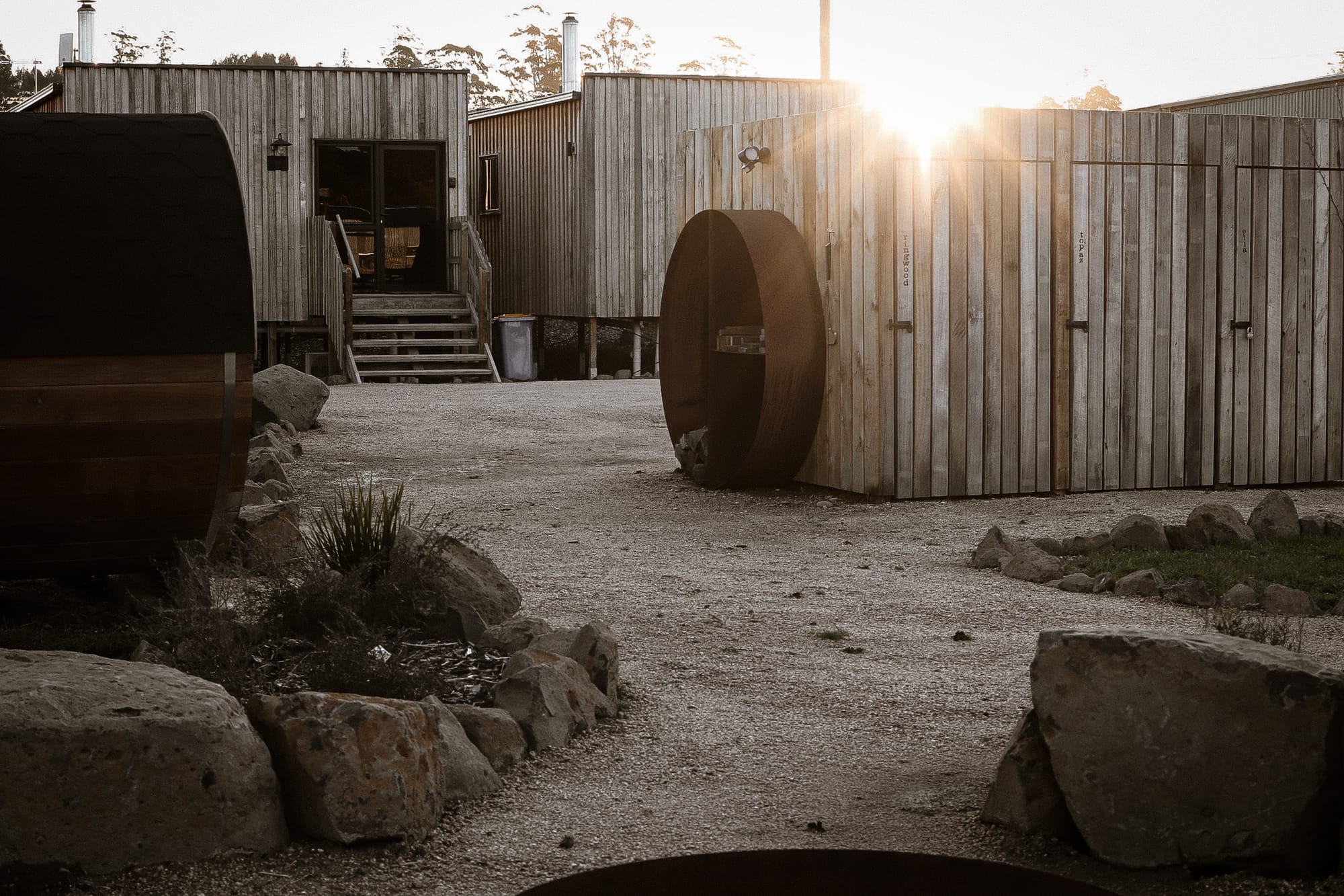 Just Down the Road. Photographer: Studio Winslow.Sunset view over a rural property with a large round wooden sauna with open doorway, positioned on a gravel ground with sparse grass and a wooden fence in the background.