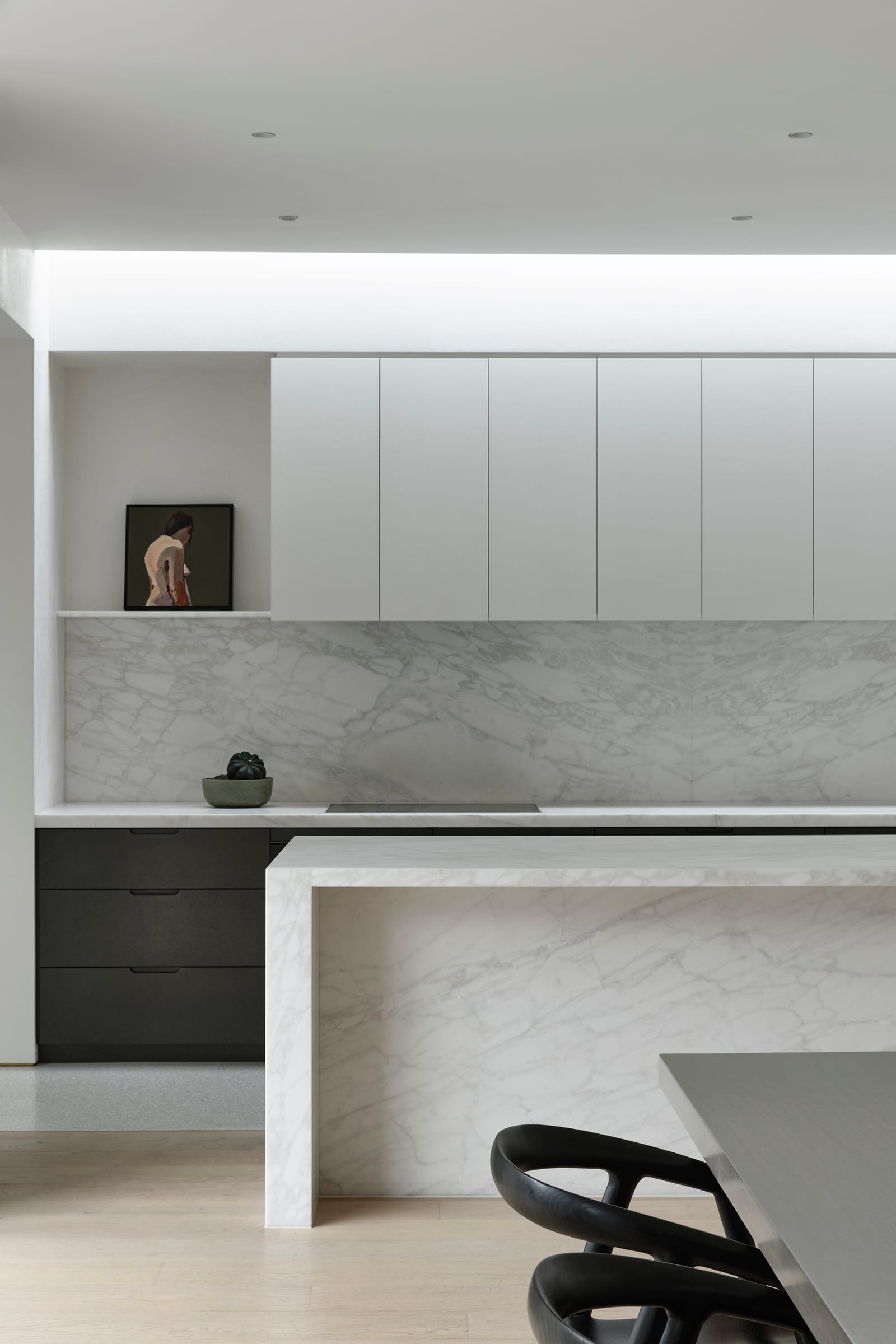 RE Residence by Inglis Architects.  interior shot focusing on a sleek, modern kitchen. The space is defined by clean lines, with a marble backsplash and countertop. White cabinetry lines the wall above the counter. A black chair with a unique, fluid design is situated at the end of the island.