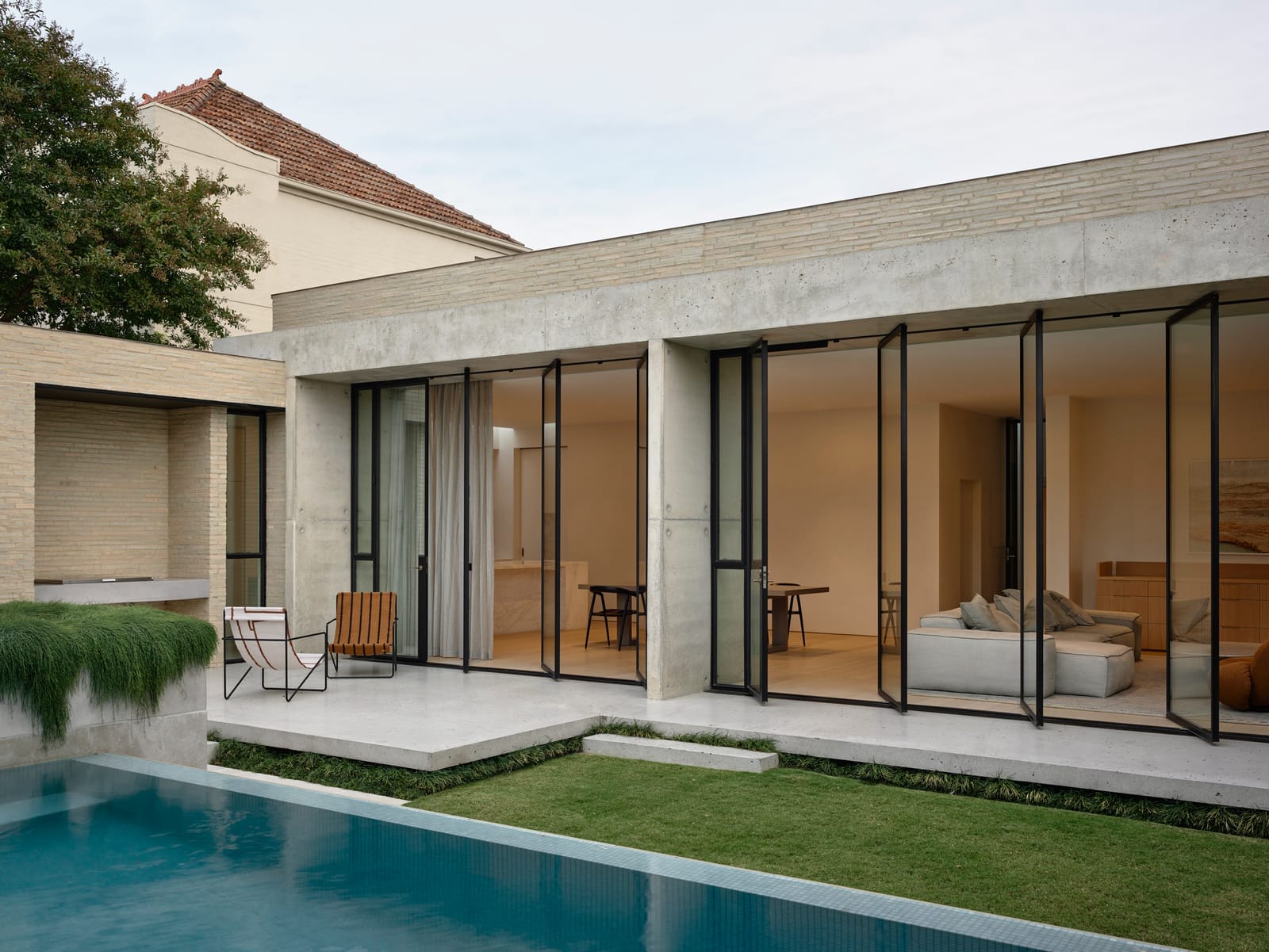 RE Residence by Inglis Architects.exterior view of a modern single-story residence with a flat roof and beige stone walls. The structure is characterized by its large glass doors and windows with slim black frames, which offer a transparent view into the interior's minimalist design. The building opens onto a patio that leads to a well-manicured lawn and a tranquil swimming pool, reflecting the clear sky and the architecture's clean lines. A wooden lounge chair sits near the pool, inviting relaxation in this serene outdoor setting.