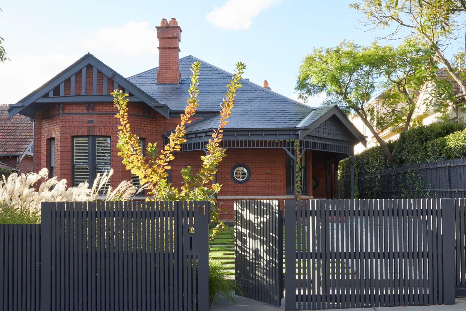 RE Residence by Inglis Architects. Edwardian style design, traditional red brick house with a tall chimney and terracotta roof tiles. The house is partially hidden behind a modern, dark metal fence and gate, with a lush garden featuring tall grasses and a bright yellow flowering plant.