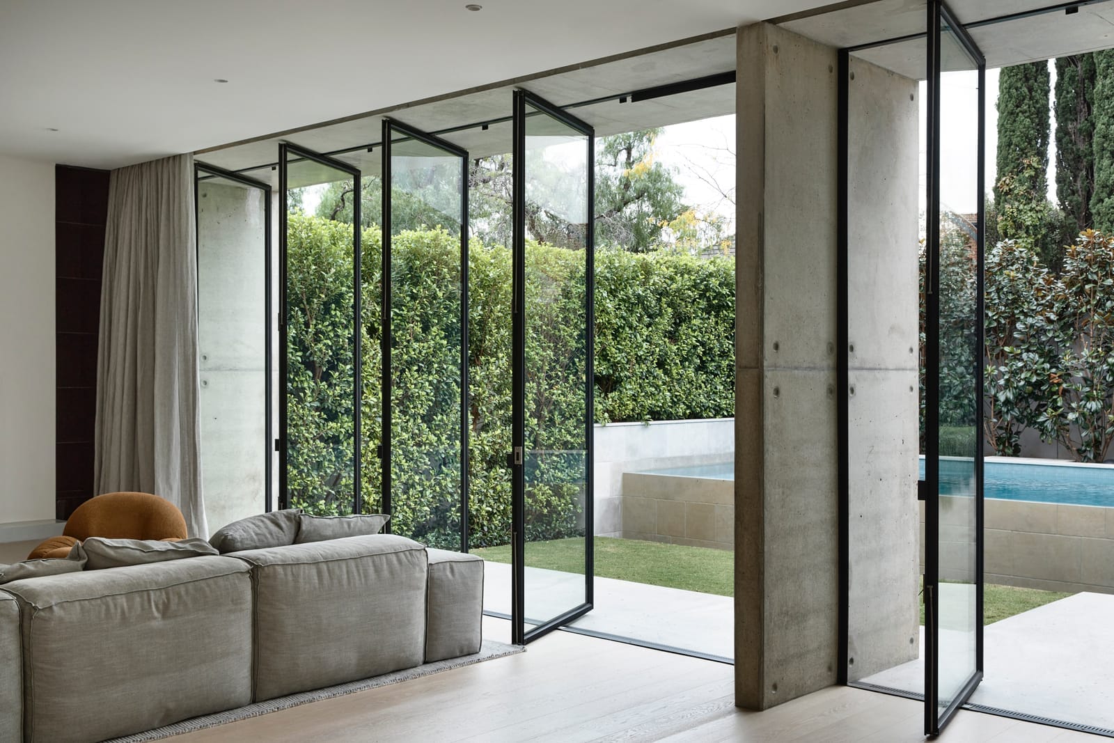 RE Residence by Inglis Architects.bright and airy corner of a living room with an open view to a garden through large glass doors with black metal frames. The doors are designed to slide away, connecting the indoor space with the outdoors seamlessly. A comfortable sofa with a light fabric is adorned with plush pillows, and a round, brown cushion sits on a modern armchair, adding a warm touch to the minimalist decor. The polished concrete floor transitions to an outdoor patio, complementing the natural green backdrop provided by the dense hedge outside.
