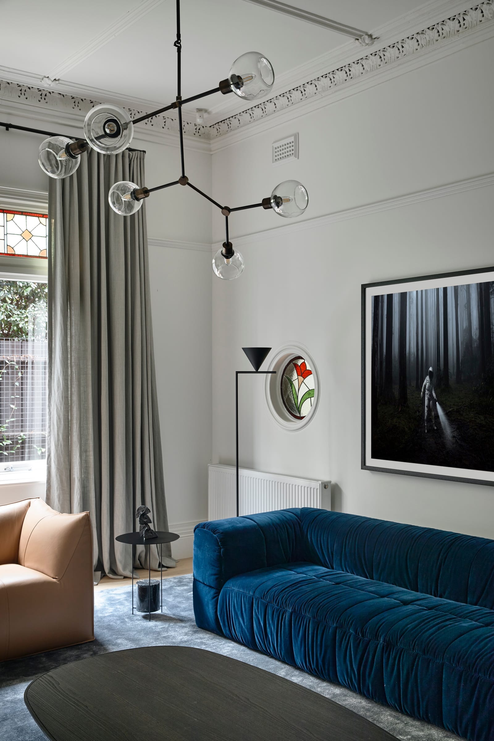 RE Residence by Inglis Architects.living room blending classic and contemporary design elements. The room features high ceilings with ornate crown moldings and a modern chandelier with multiple spherical bulbs on branching metal arms. A deep blue velvet sofa provides a striking contrast against the neutral-toned walls. Sheer curtains drape elegantly beside a window, and a large framed photograph of a misty forest adds an artistic touch. A geometric sculpture and a black floor lamp contribute to the room’s sophisticated aesthetic.