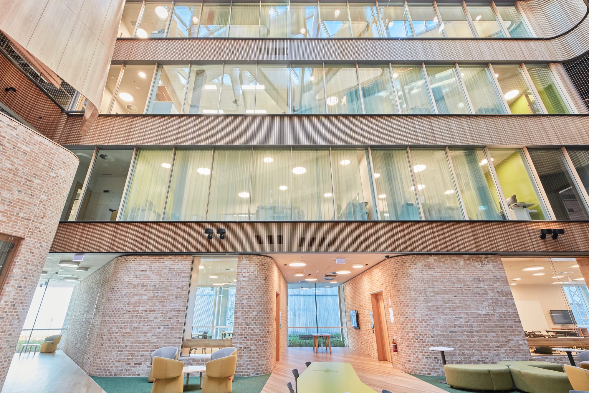 Tasmanian Timber: River's Edge. Photography: Dave Groves.An upward perspective of a multi-level building's interior, highlighting the juxtaposition of rustic brick walls and contemporary glass windows. The open space is well-lit with natural light, and the mezzanine floors with glass balustrades contribute to the open-concept design. The area seems to function as a communal space with seating areas visible on the ground level.
