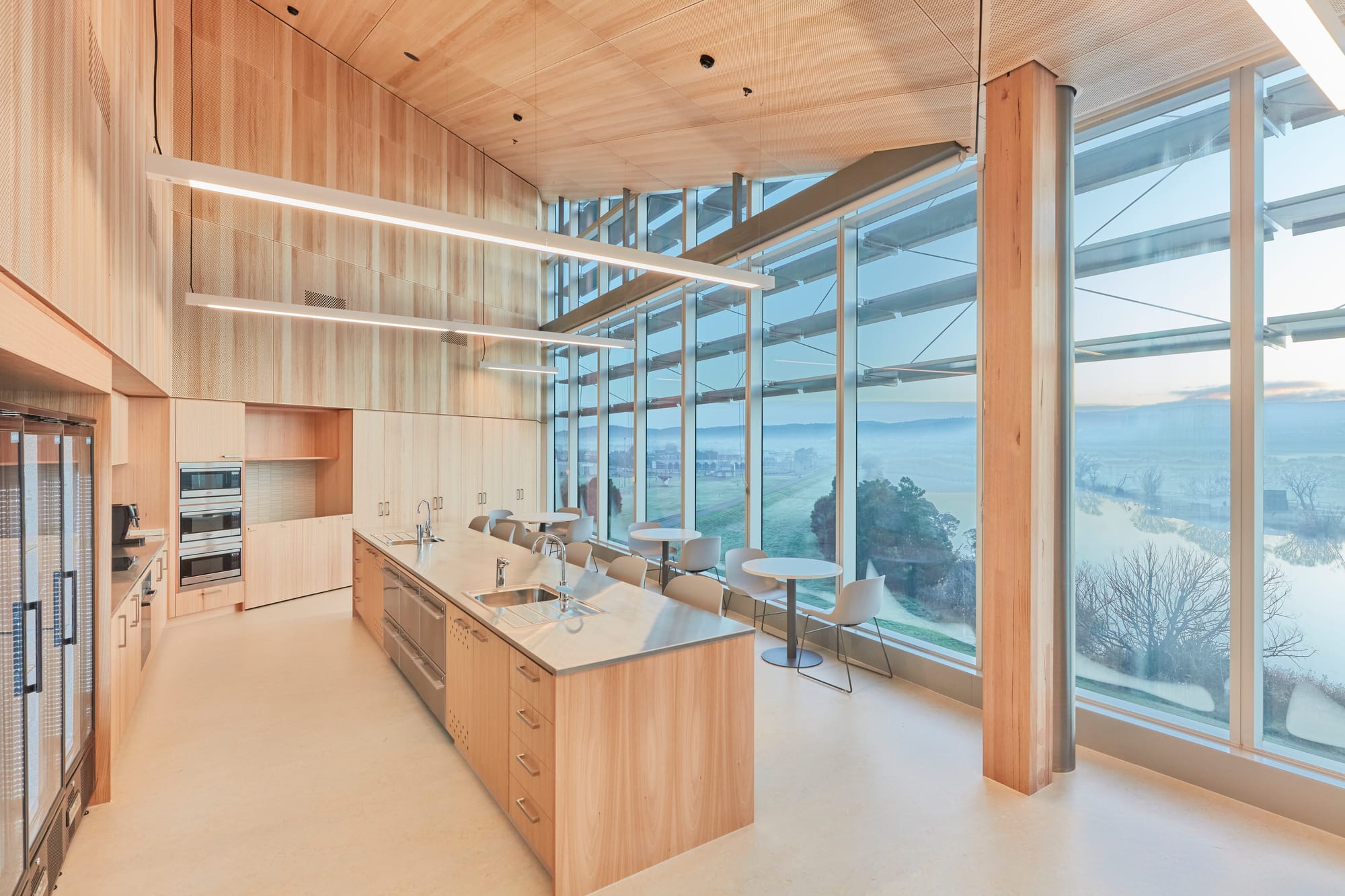 A bright and airy dining or meeting area with a wall of large windows offering a sweeping view of a scenic landscape with a clear sky. The room features a light wooden floor and is furnished with sleek, modern chairs and round tables. The interior wall is clad in warm, light-colored wood panels, and the overall ambiance suggests a calm and inviting space for socializing or working.