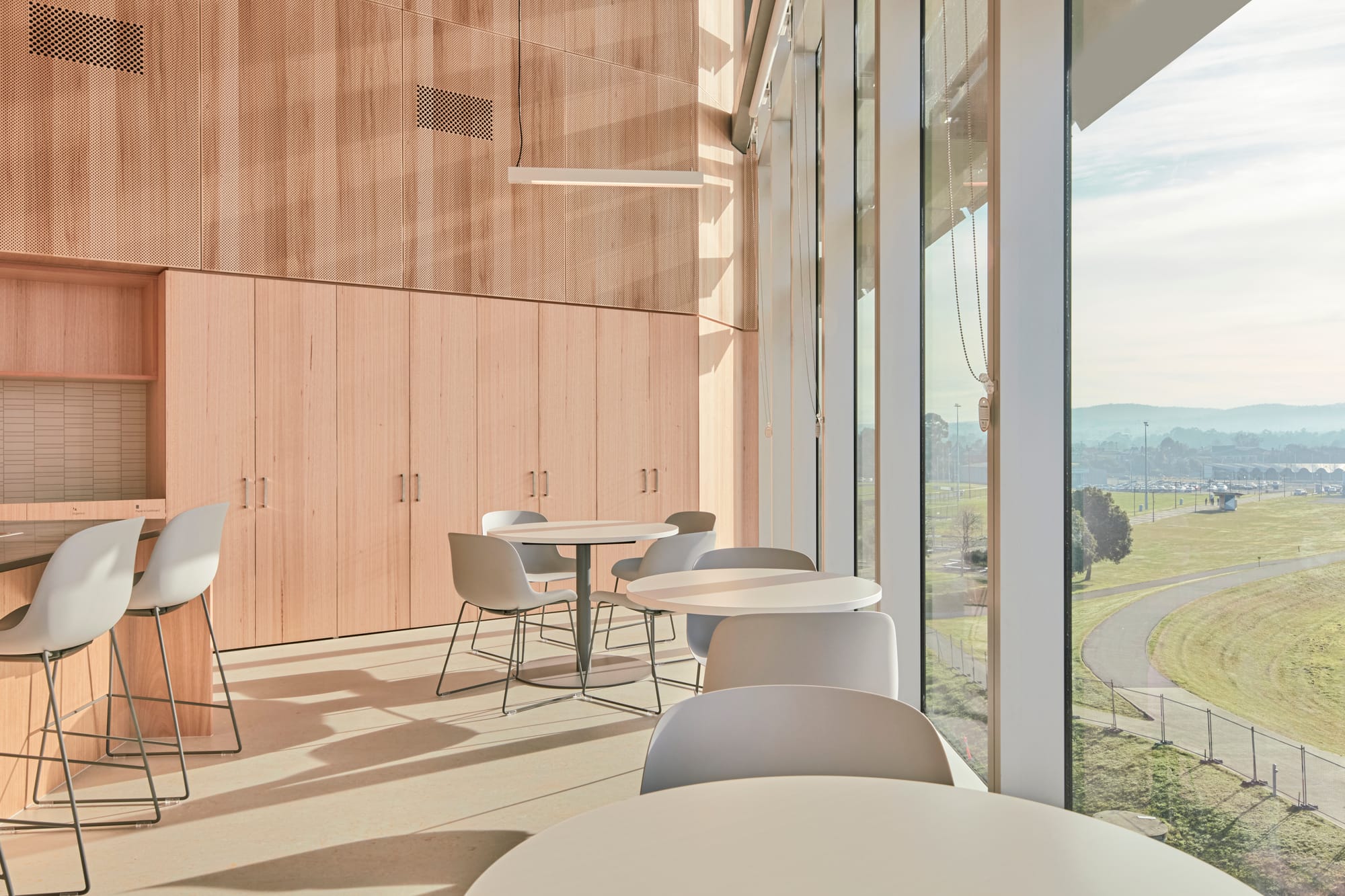 Tasmanian Timber: River's Edge. Photography: Dave Groves.A bright and airy dining or meeting area with a wall of large windows offering a sweeping view of a scenic landscape with a clear sky. The room features a light wooden floor and is furnished with sleek, modern chairs and round tables. The interior wall is clad in warm, light-colored wood panels, and the overall ambiance suggests a calm and inviting space for socializing or working.