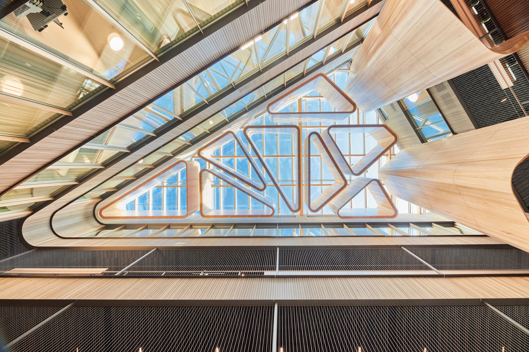 Tasmanian Timber: River's Edge. Photography: Dave Groves.n interior view of a modern atrium from a low angle, showcasing a geometrically complex glass ceiling with wooden frames. The design creates a striking pattern of triangles and trapezoids with the sky visible through the glass. The sides of the atrium feature multiple levels with wooden and metal railings, emphasizing the building's verticality and the use of natural materials.