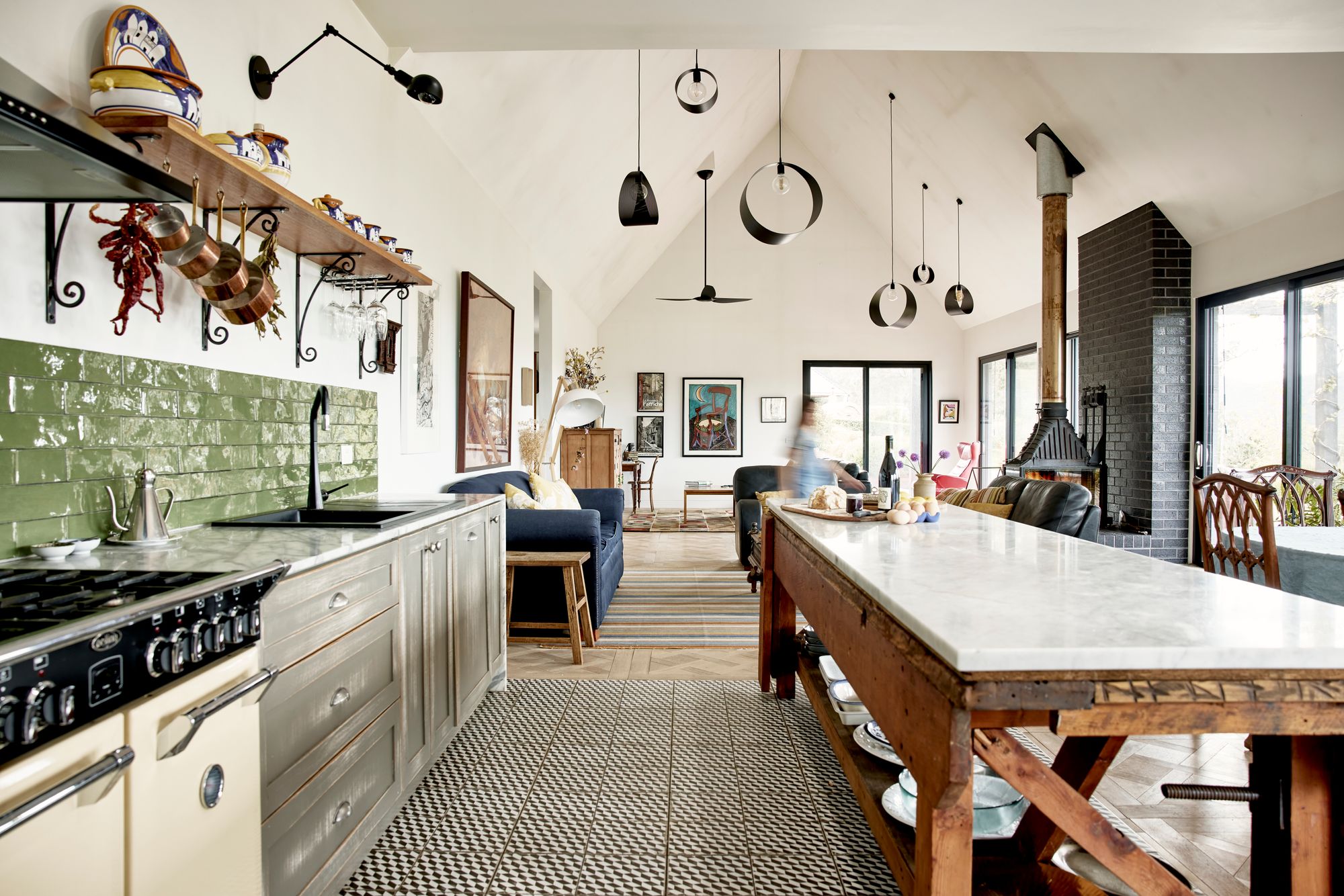 well-lit kitchen extending into a living area, characterized by a high vaulted ceiling and multiple hanging pendant lights. The kitchen area features a large island with a marble countertop, grey cabinetry, and a green tile backsplash. There are open shelves holding various kitchen items and utensils. A professional-grade stove and sink with a modern faucet are visible. The living area includes a fireplace with a black chimney, comfortable seating, and a striped area rug. Artwork and decorative items can be seen throughout the space, providing a homely atmosphere. Large windows allow for ample natural light. In the background, a person appears to be in motion, contributing to the lived-in feel of the space.