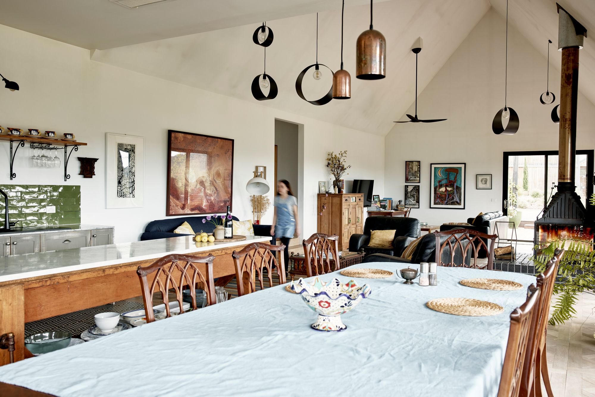 The kitchen has a marble-topped island, green tiled backsplash, and wooden shelves with kitchenware. Various pendant lights hang above the dining table. Artworks and comfortable furnishings decorate the lounge. A person in motion adds liveliness to the scene, illuminated by ample natural light.