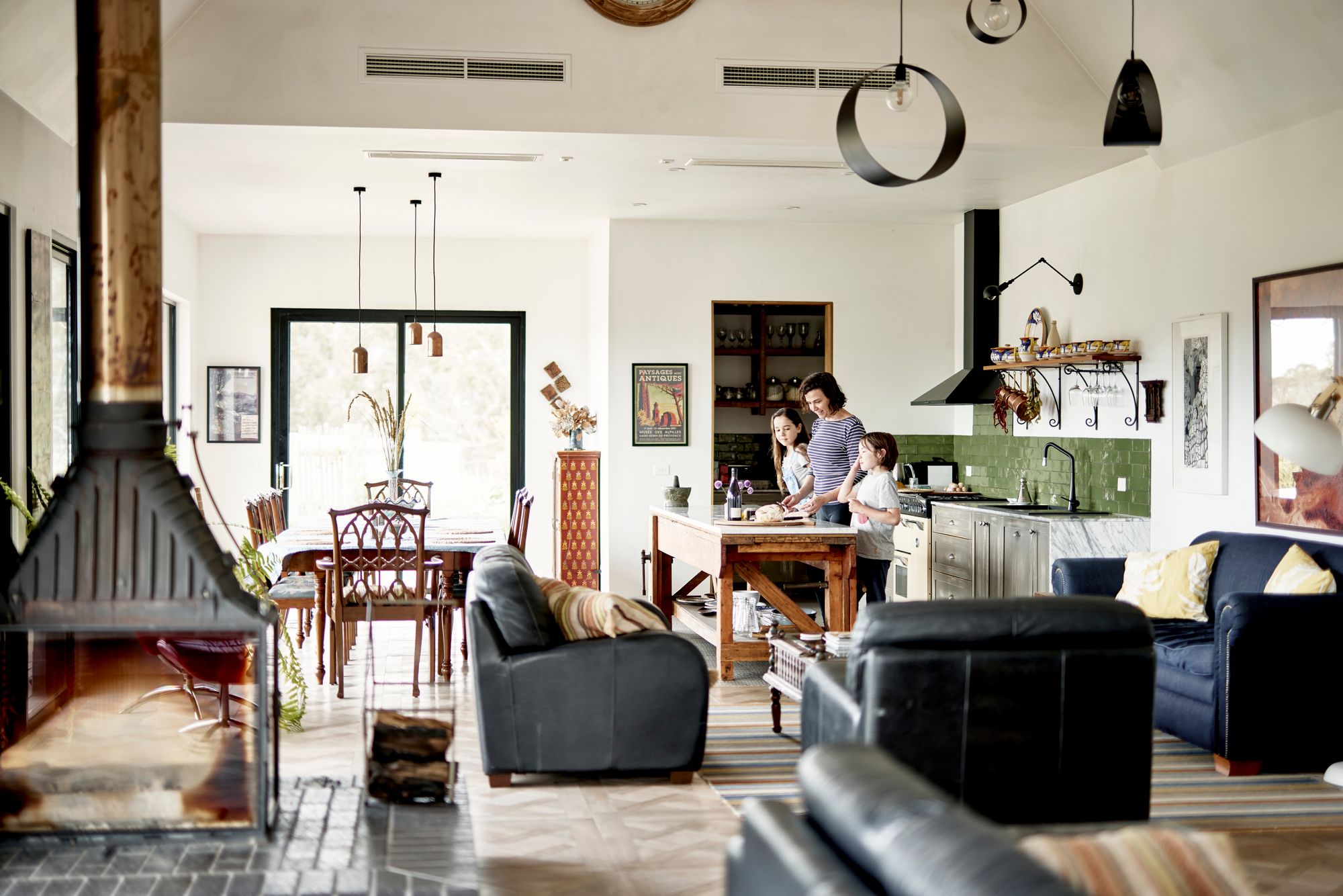 The Glut Farm. Photography copyright of The Glut Farm. homey and vibrant open-plan living space with a family engaged in an activity in the kitchen area. The kitchen has a green tile backsplash and is equipped with stainless steel appliances and a central island. The dining area includes a wooden table set with chairs, and the living area has comfortable leather seating. The interior is well-lit with natural light streaming in through large windows, and the décor includes various artworks and plants, creating a welcoming atmosphere.