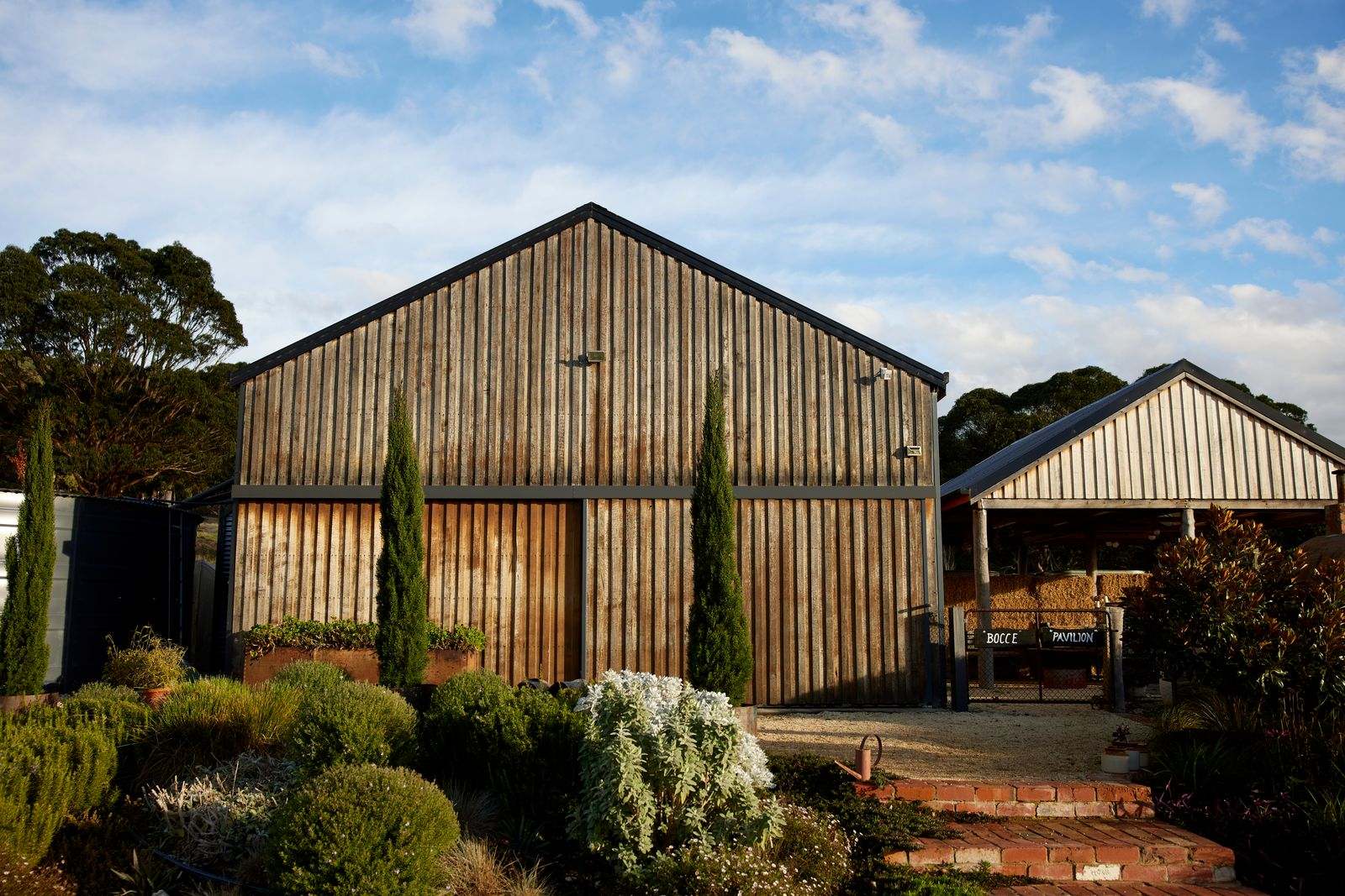 The Glut Farm. Photography copyright of The Glut Farm. rustic barn-style building with weathered wooden siding and a prominent gabled roof. It features tall, narrow windows and is surrounded by well-maintained landscaping with shrubs and trees.
