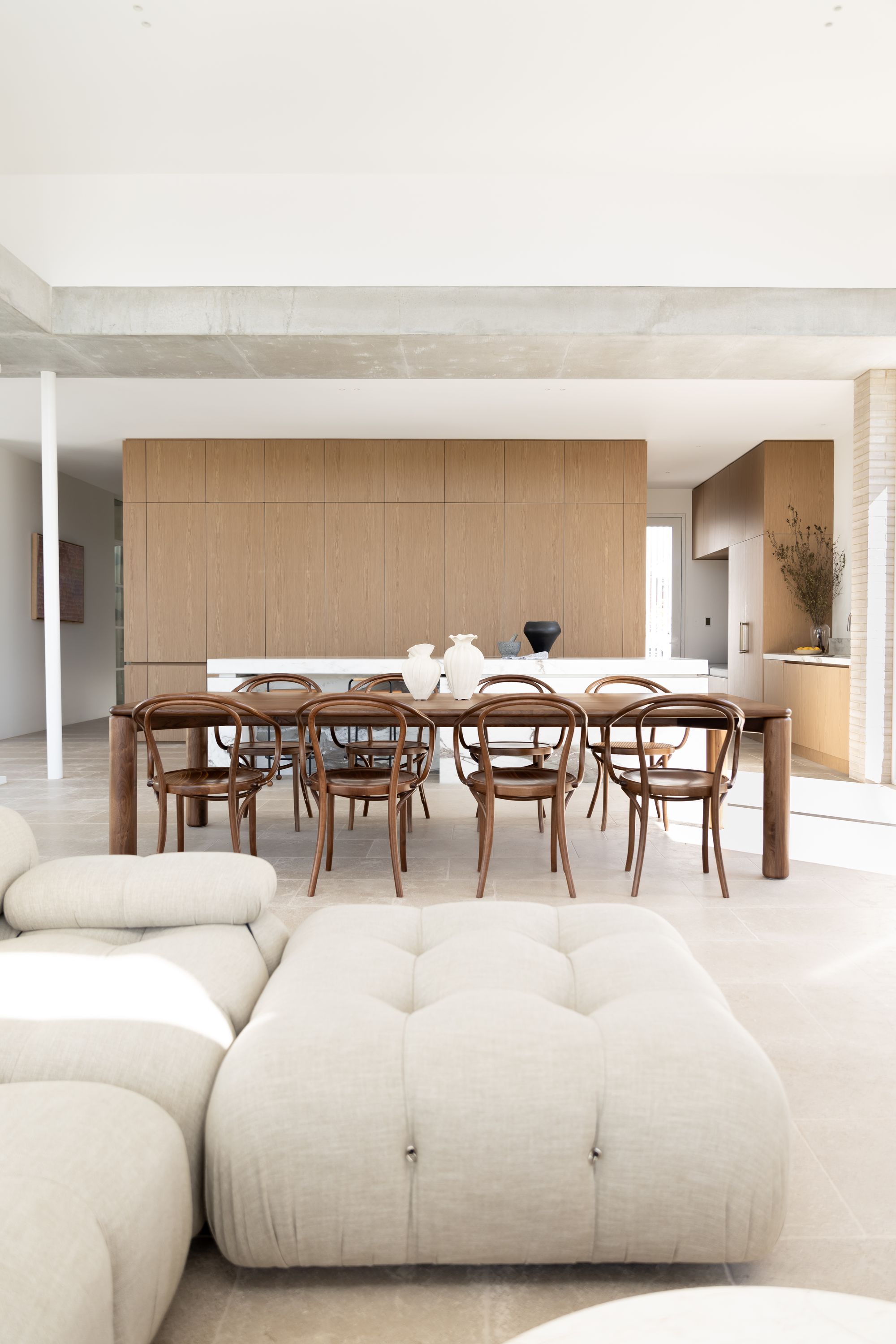 Timberwolf Design: Ladovi Table. modern and minimalist dining area featuring a large wooden dining table surrounded by bentwood chairs. The room has a neutral color palette with beige tiled flooring, wood paneling on the walls, and a white ceiling. The natural light fills the space, giving it an airy feel. The furniture arrangement and the choice of materials suggest a contemporary design with a nod to classic elements.