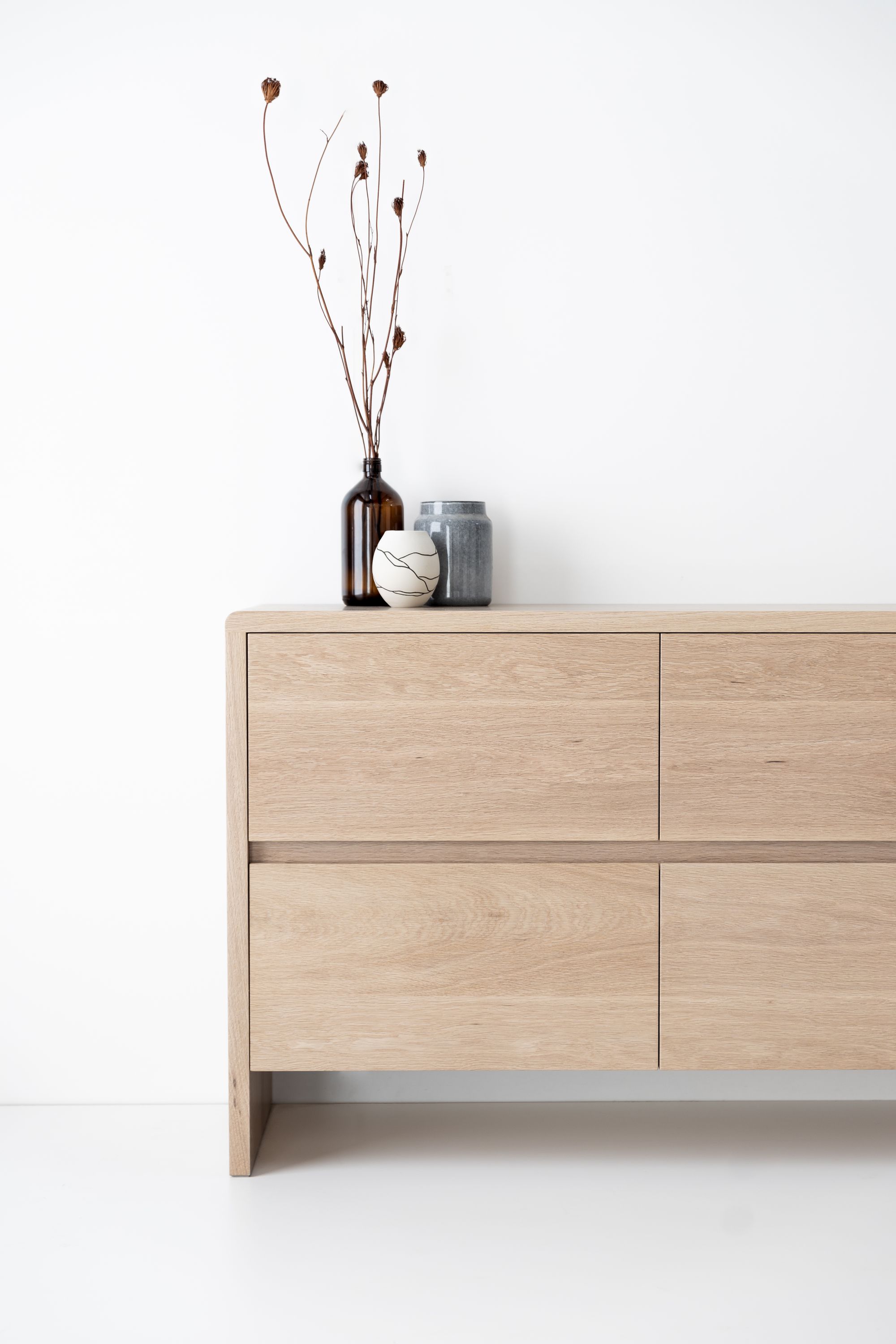 Timberwolf Design: Sova Credenza in American Oak Natural.modern wooden sideboard with a smooth finish. The sideboard is styled with a tall brown glass bottle holding dried plants, a grey pottery piece, and a small cup with a cracked pattern. The simple arrangement and natural materials give the scene a serene and organic feel, while the white wall behind ensures that the focus remains on the sideboard and its decor.