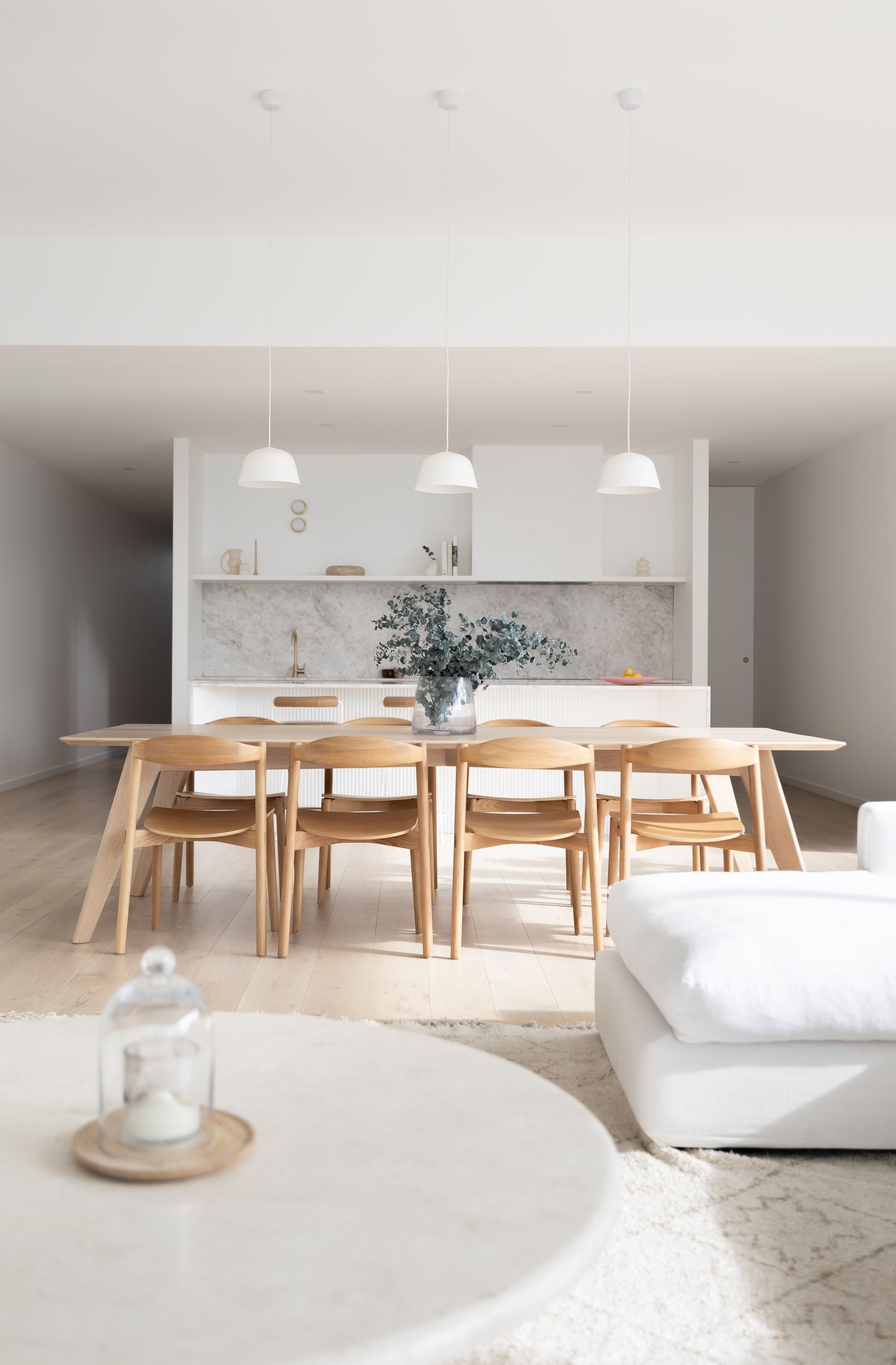 Timberwolf Design: Elk Dining Table.The dining space is defined by a light wood table and matching chairs on a textured round rug, highlighting the warm tones of the wooden floor. Above the table hang two simple white pendant lights. A glass vase with blue foliage serves as a centerpiece, and the background reveals a kitchen counter with a marble backsplash, creating a clean and sophisticated atmosphere.