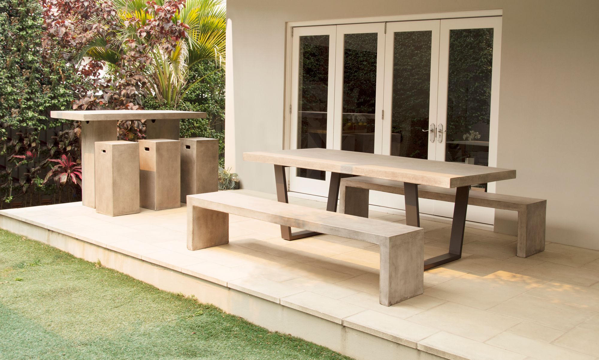 Remarkable Outdoor Living: Zen Outdoor Dining table with Aluminium Trapezoid shade Leg. A contemporary outdoor dining set, featuring a concrete table and benches, is set against a lush garden backdrop, offering a harmonious blend of man-made structure and natural beauty.