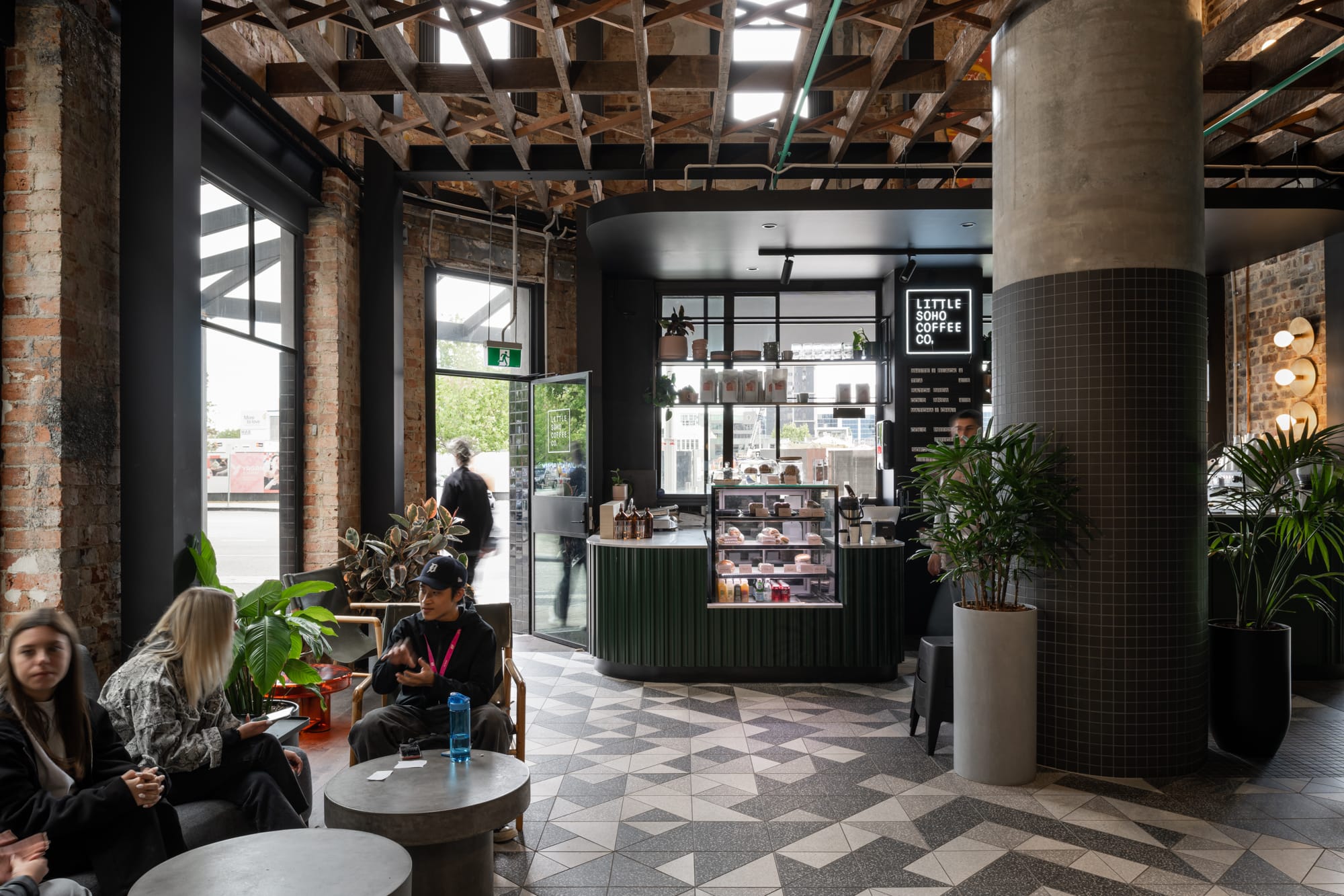 Thinking Differently About a Home Gives You Affordable ChoicesA spacious coffee shop with an industrial vibe, featuring exposed brick walls and large windows. People are seated at tables, enjoying the ambiance. A central column is wrapped with a tiled counter, and the shop extends into a light-filled area with a display case.