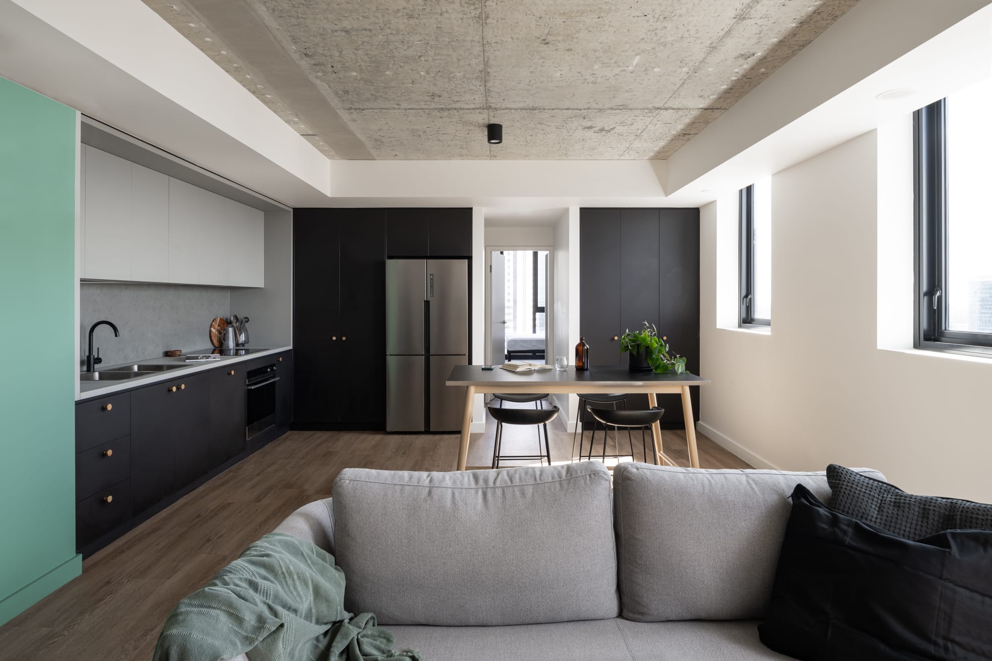 Thinking Differently About a Home Gives You Affordable Choices. A modern apartment interior with a dark kitchen and a central island. The open plan layout connects the kitchen to a cozy living area with a gray sofa, all under a white ceiling with recessed lighting.
