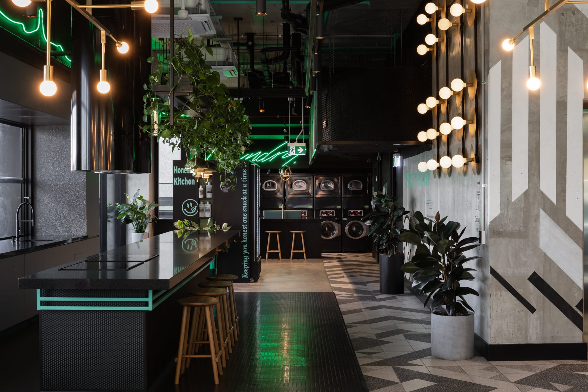 Thinking Differently About a Home Gives You Affordable ChoicesAn industrial-style kitchen bar with dark green accents and hanging Edison bulbs. The bar features a mirrored backsplash with a green neon sign. Lush plants add a touch of nature to the space, which is balanced by concrete floors and a patterned tile wall.