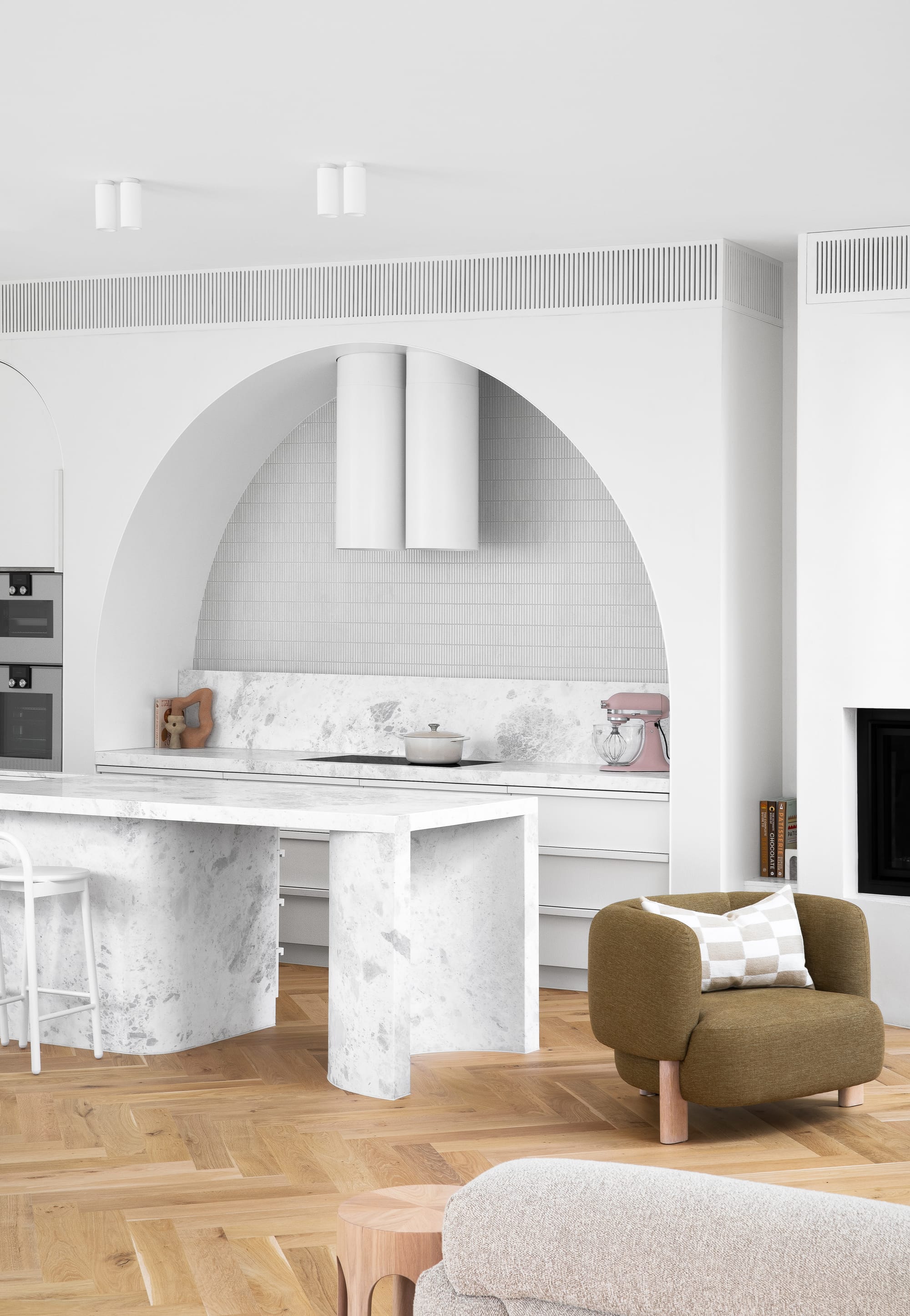 The Grove by Taouk Architects.A modern and spacious kitchen and living area with white walls and high ceilings. The kitchen features white marble countertops, a central island, and a backsplash with an arched niche above the stove. The open-plan area includes a living space with a comfortable tan sofa and armchair, with natural hardwood flooring throughout. The room is illuminated by natural light, creating a welcoming and serene ambiance.