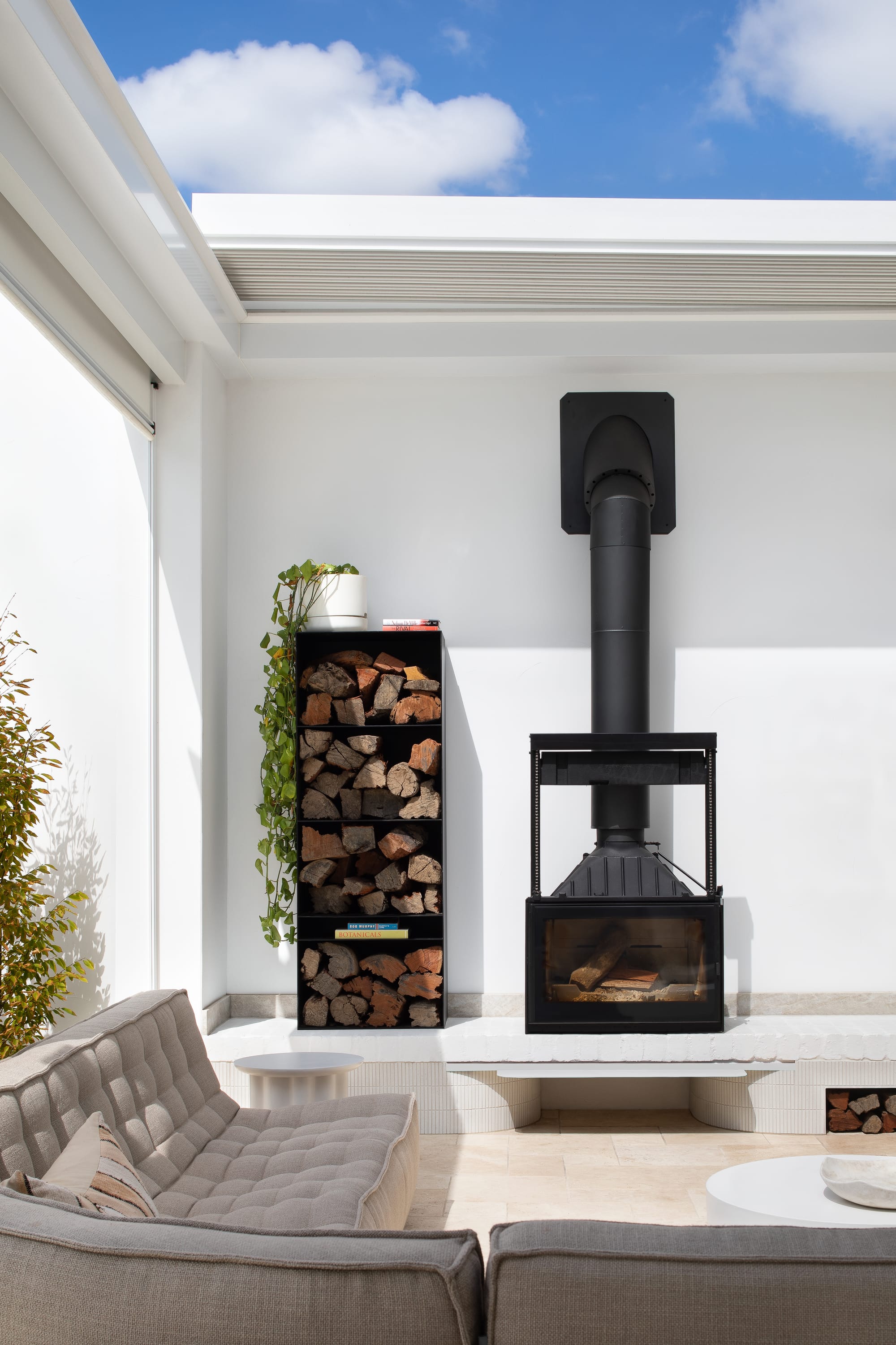 The Grove by Taouk Architects.A cozy indoor nook featuring a contemporary freestanding black fireplace with a metal flue that extends up to a high white ceiling. To the side of the fireplace, a neat stack of firewood is artfully arranged, adding a rustic touch to the modern space. The area is furnished with a comfortable gray sofa, throw pillows, and a low-profile coffee table, creating a warm and inviting space to relax.