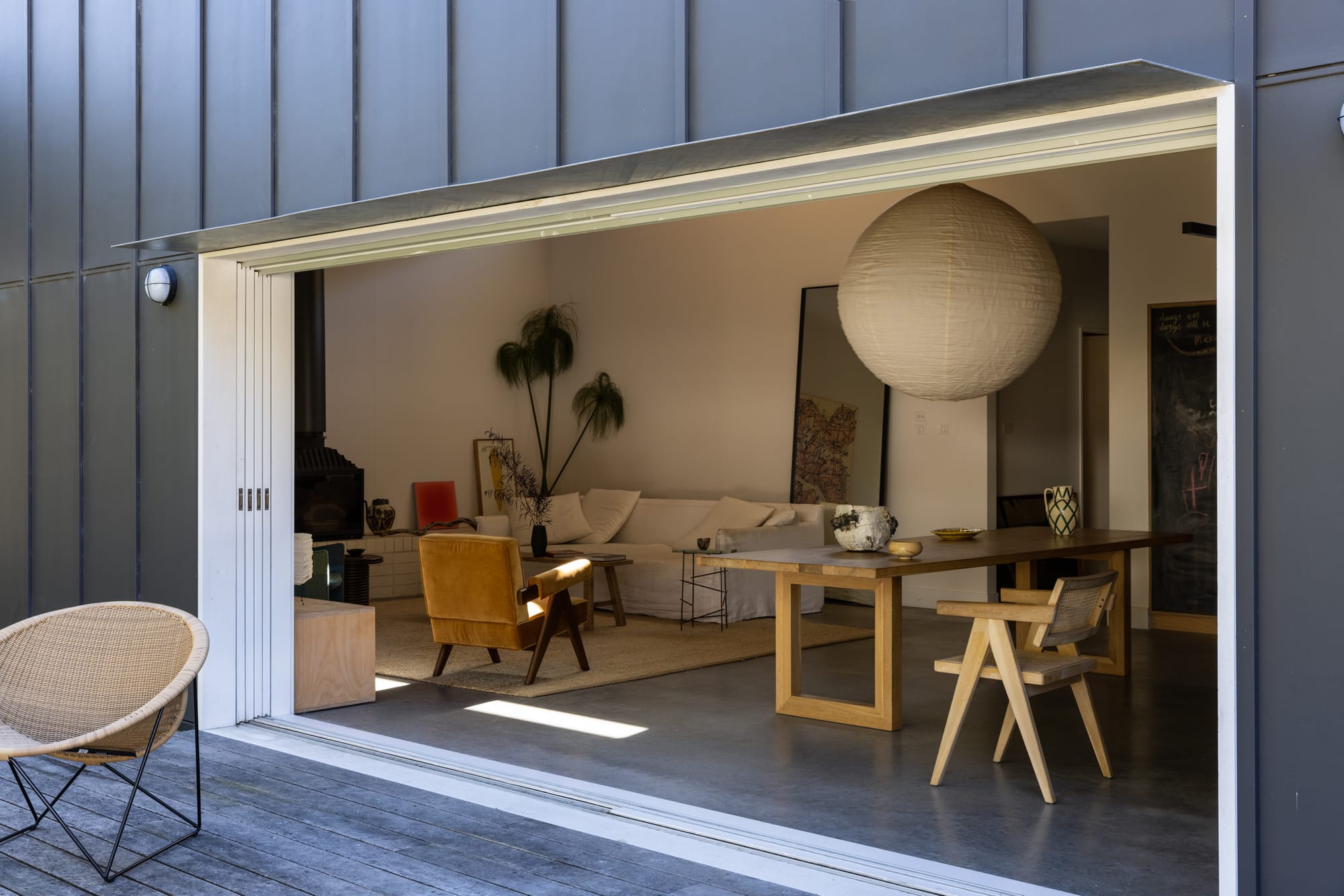 Drip-Dry House by Marker Architect. Wide angle image showing the interior and exterior view through a large, sliding door recess. Exterior of facade on right hand side of image shows black timber paneled walls and weathered timber deck. Interior of space depicts cosy and simple living space featuring a white couch, mustard yellow armchair and jute rug to the right and a long timber dining table to the right. A large, white spherical pendant hangs over concrete floors.