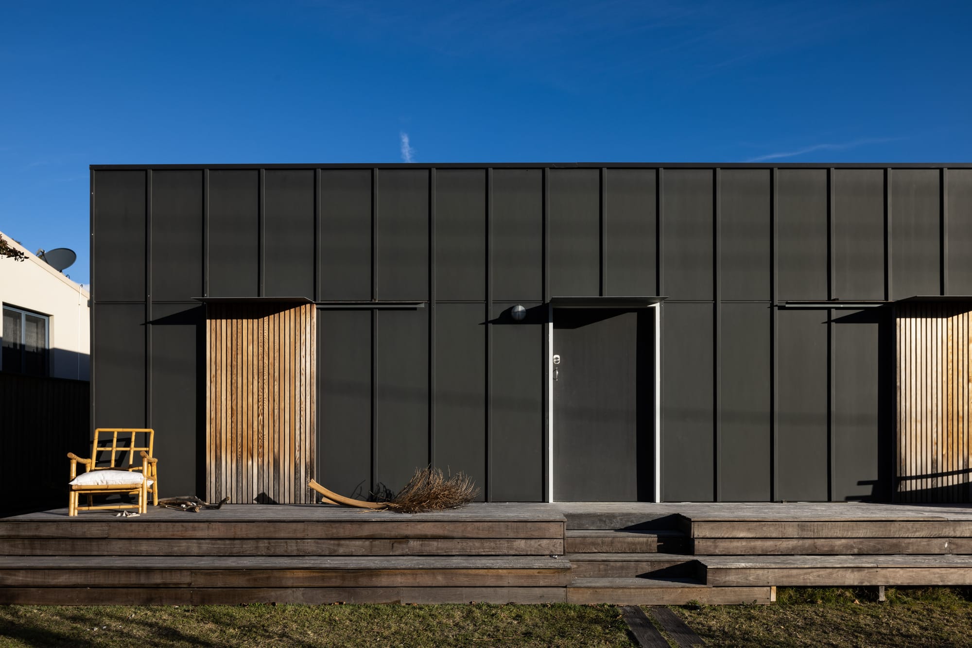 Drip-Dry House by Marker Architect. Exterior facade of residential building, clad in black timber panels in simple, rectangular shape. Tiered, weathered timber deck extends forwards from facade in front of a plain, black windowless door. Timber clad panels conceal windows. Cloudless blue sky above.
