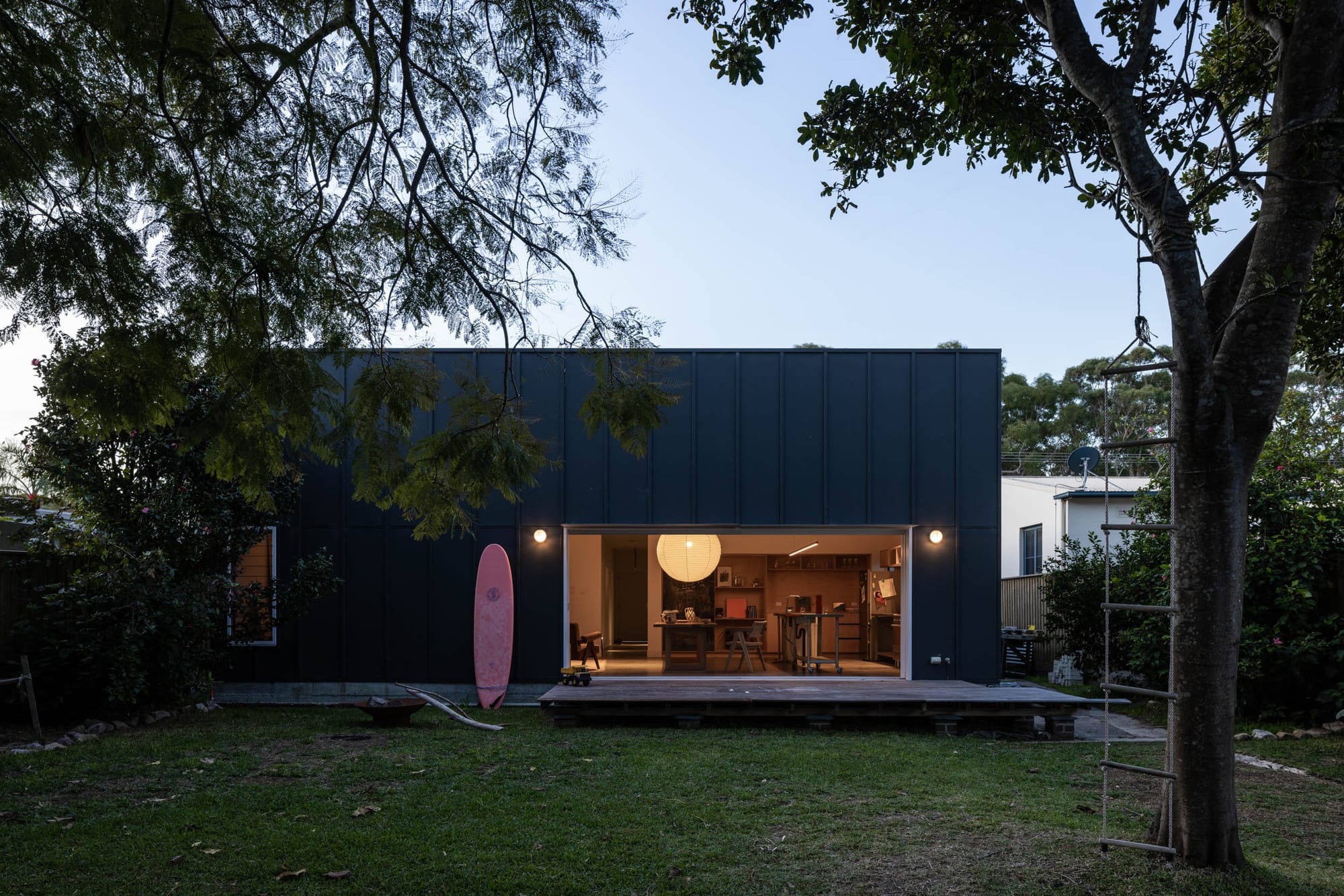 Drip-Dry House by Marker Architecture & Design. Rear elevation of the beach house showing a red surfboard leaning against the real wall and a glimpse into the dining and kitchen areas with warm yellow light coming from the pendant light