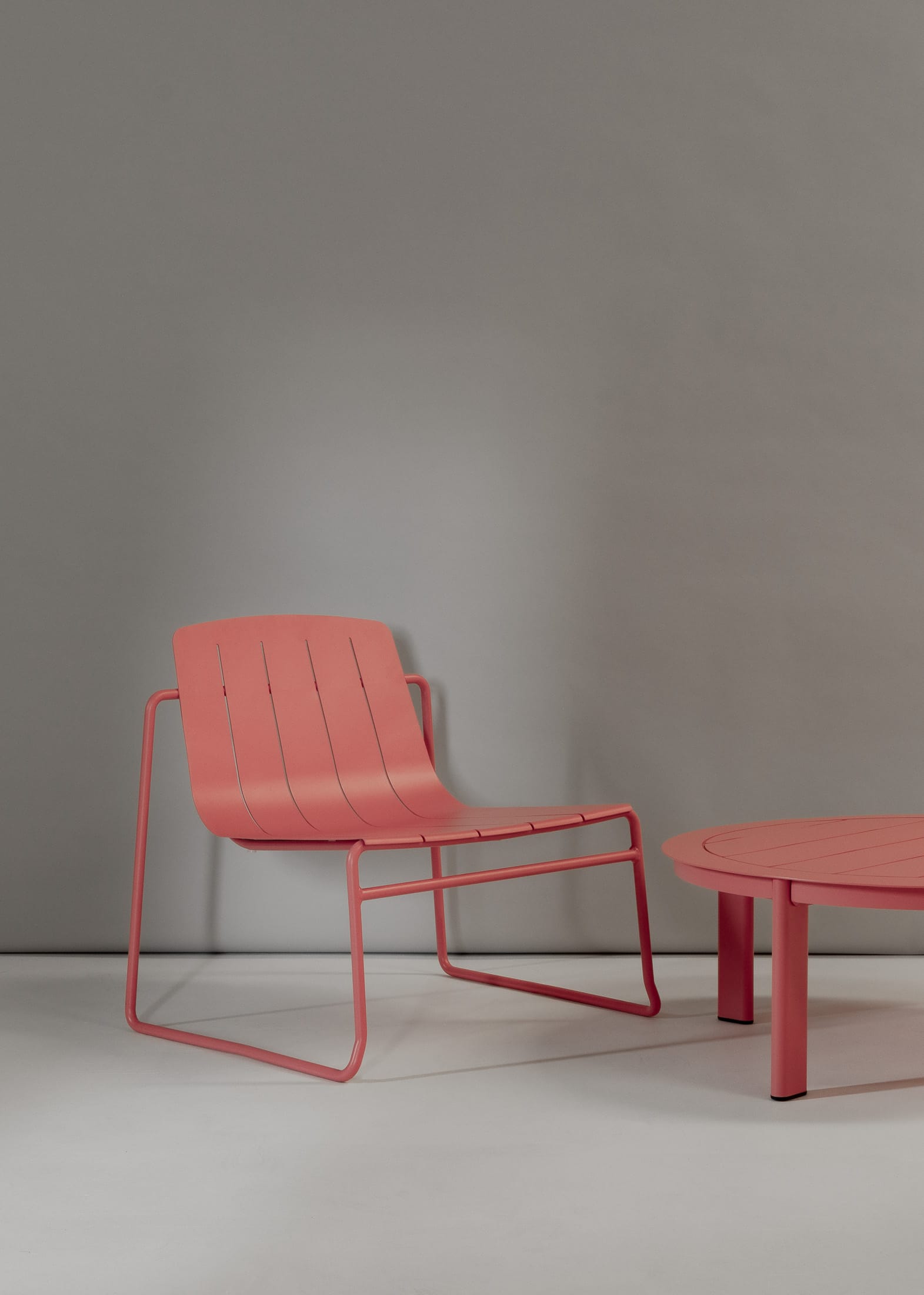 Franka Outdoor: Harper Outdoor Coffee Table & Frankie Outdoor Lounge Chair in Melon. contemporary outdoor lounge area featuring a single pink metal chair with broad armrests and a low back, accompanied by a round, flat-topped pink side table, set against a simple grey backdrop.