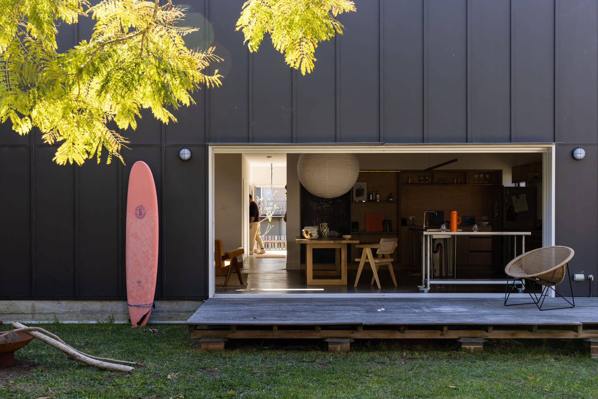 Drip-Dry House by Marker Architect. Wide angle shot of a residential exterior rear-facade. Black timber double story walls in a simple industrial style. Weathered deck extends from large opening in facade. Minimal living space visible through opening. Red surfboard leaning against exterior wall. Green grass in foreground of image.