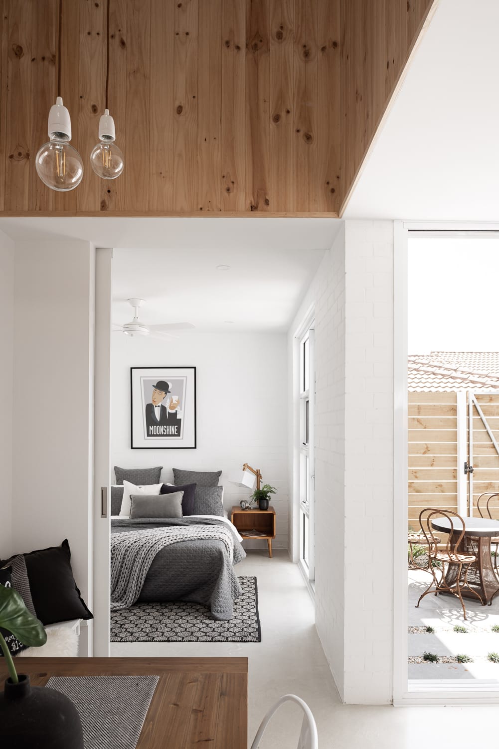 This image showcases a modern bedroom with a view into an outdoor seating area. The bedroom features a cozy gray bed with a knitted throw and patterned pillows, set against a simple white painted brick wall. Above the bed hangs a framed "MOONSHINE" poster, adding a touch of personality to the room. To the side of the bed, there's a wooden nightstand with a plant, enhancing the room's natural feel. The ceiling above is lined with warm wooden panels, and two simple pendant lights dangle, casting a soft glow. Through the open sliding glass door, one can see a quaint outdoor area with a wooden table and chairs set upon a pebbled patio, inviting relaxation.