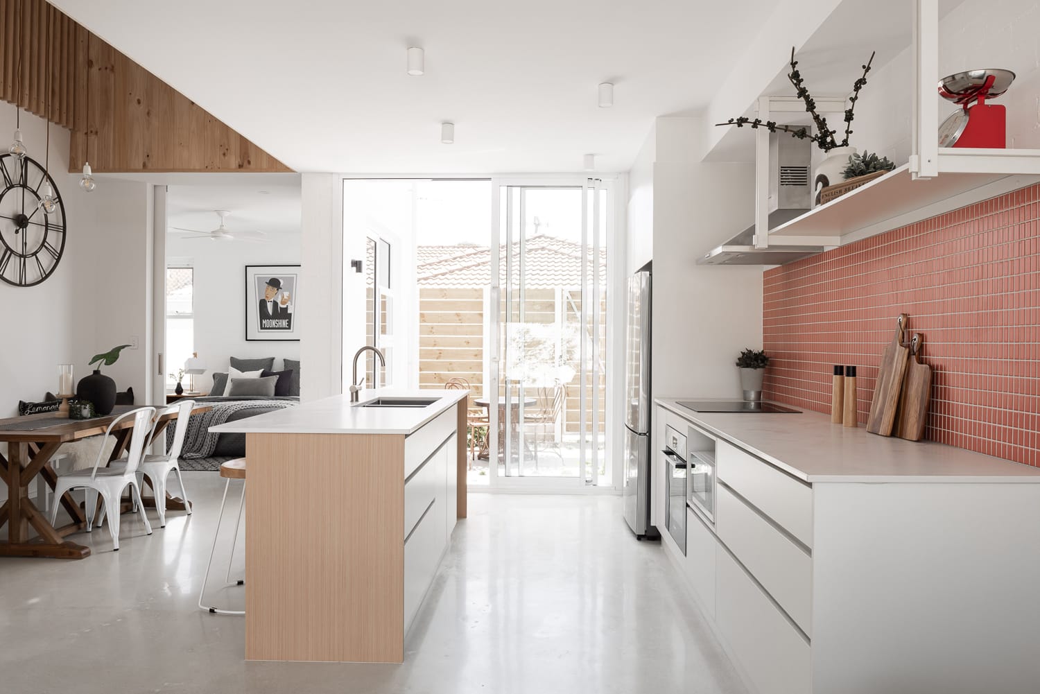 Carrington Street Terraces by MDC Architects.The interior view highlights a bright, open-plan kitchen and dining space with sleek, contemporary design. The kitchen boasts white countertops and cabinetry, with a central island featuring a wooden countertop. A pink-tiled backsplash adds a pop of color. The dining area includes a wooden table with modern chairs and shelving adorned with decorative items. Natural light floods in from the large windows and door at the back.