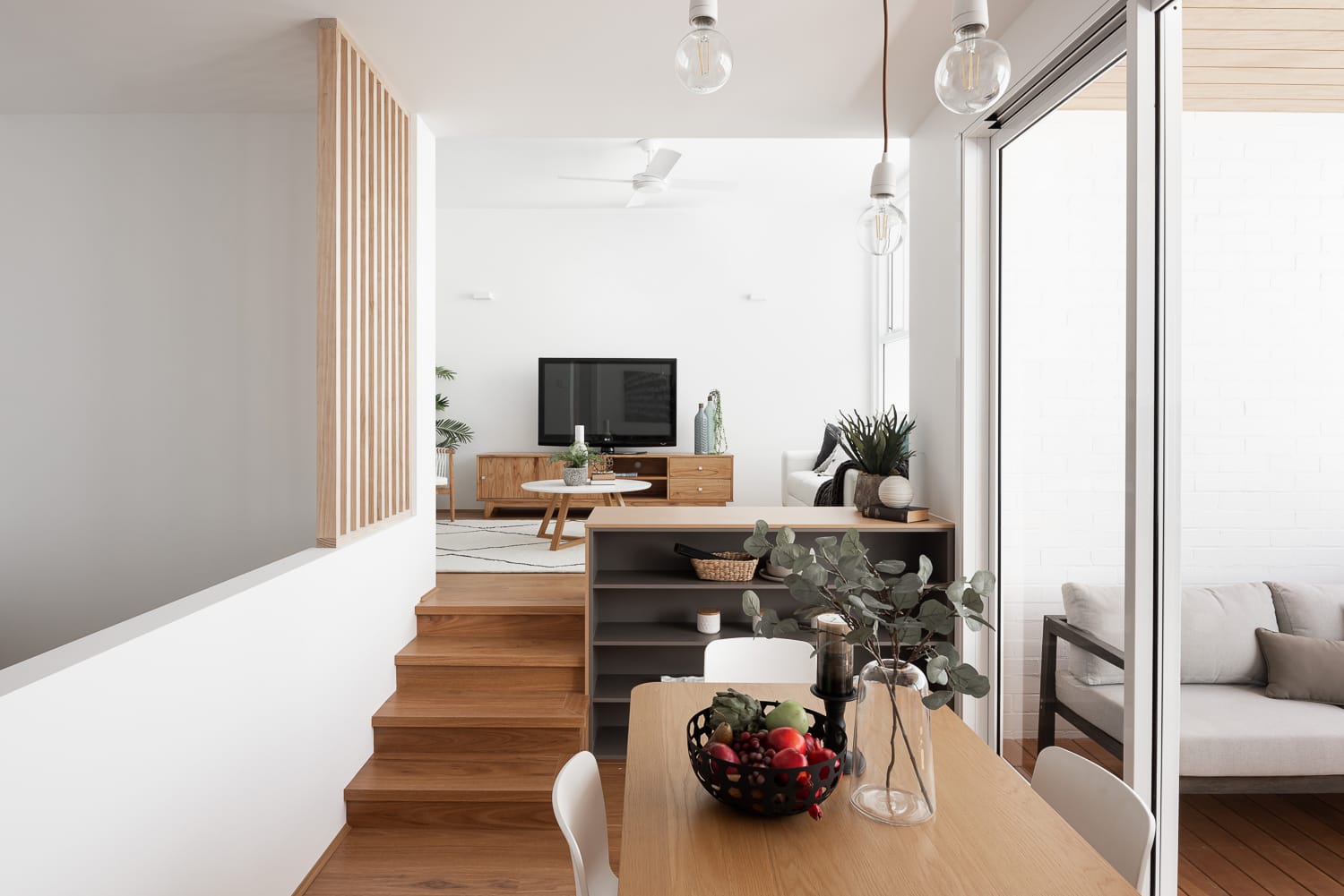 Carrington Street Terraces by MDC Architects.This image features a modern, split-level interior with a dining area in the foreground and a lounge space a few steps up. The dining area includes a simple white table with chairs and a large clear vase with eucalyptus branches. Pendant lights hang above, and natural light streams in through large sliding glass doors. The lounge area is minimalistic with a wooden media console, flat-screen TV, and a cozy light gray sofa with cushions. The white walls and wooden flooring create a warm, inviting atmosphere, enhanced by indoor plants, adding a touch of greenery to the space.