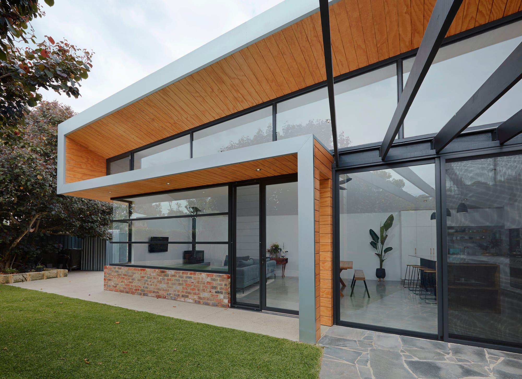 Sharp House by Steelehouse Architecture.his image shows the exterior of a modern home featuring a unique blend of materials, with exposed brickwork, wooden cladding, and large glass windows. The architecture showcases clean lines and geometric forms, with a cantilevered upper section creating a covered outdoor area. The garden is well-kept, with a stone pathway leading to the entrance.