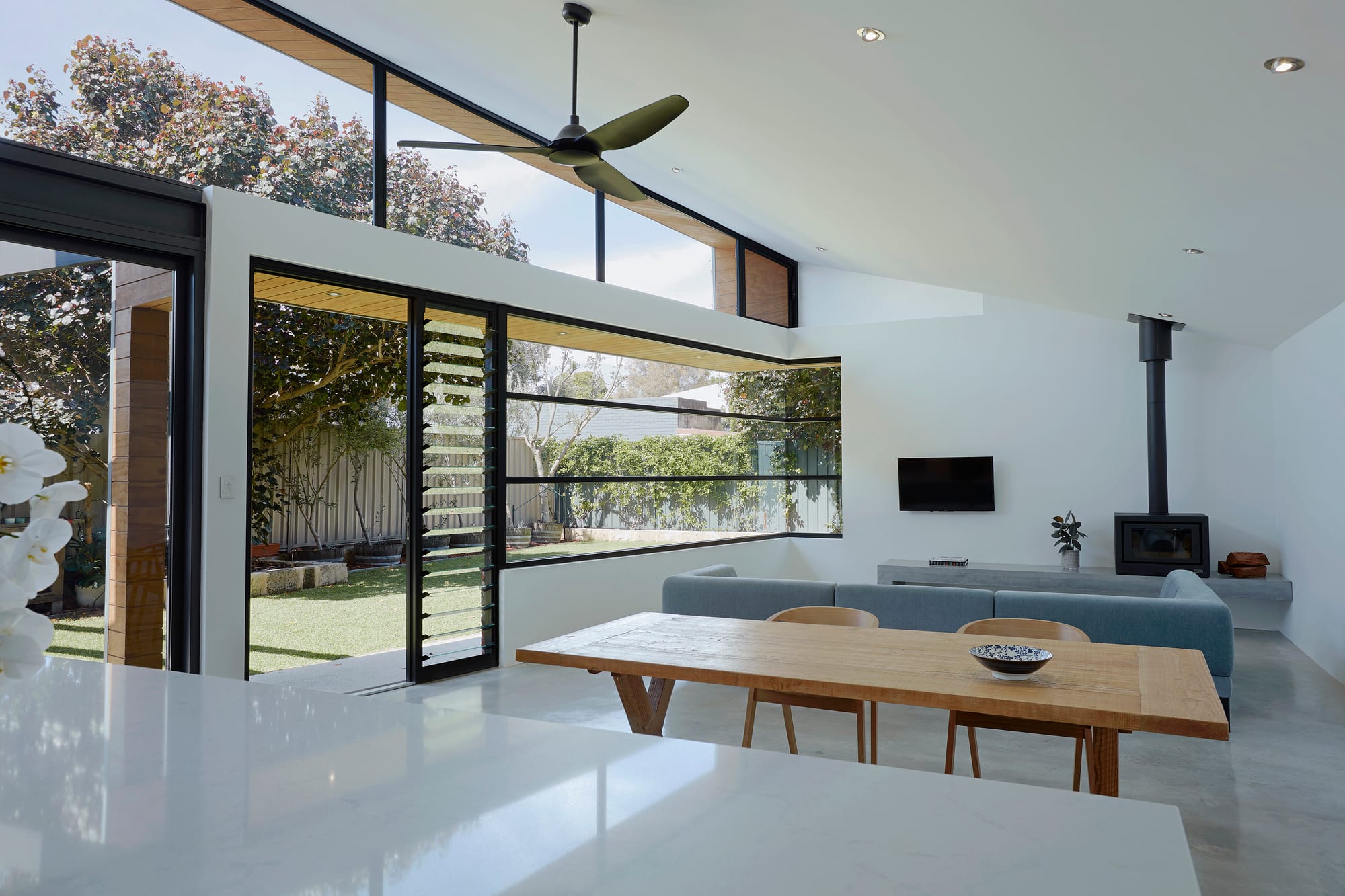 Sharp House by Steelehouse Architecture.The photo depicts a contemporary, spacious kitchen and dining area with a minimalist design. The kitchen features sleek white cabinetry with a contrasting dark backsplash and a large kitchen island with a white marble countertop. The dining area includes a solid wood table with matching chairs. Large windows and sliding doors allow for an abundance of natural light and a seamless connection to the outdoor garden.