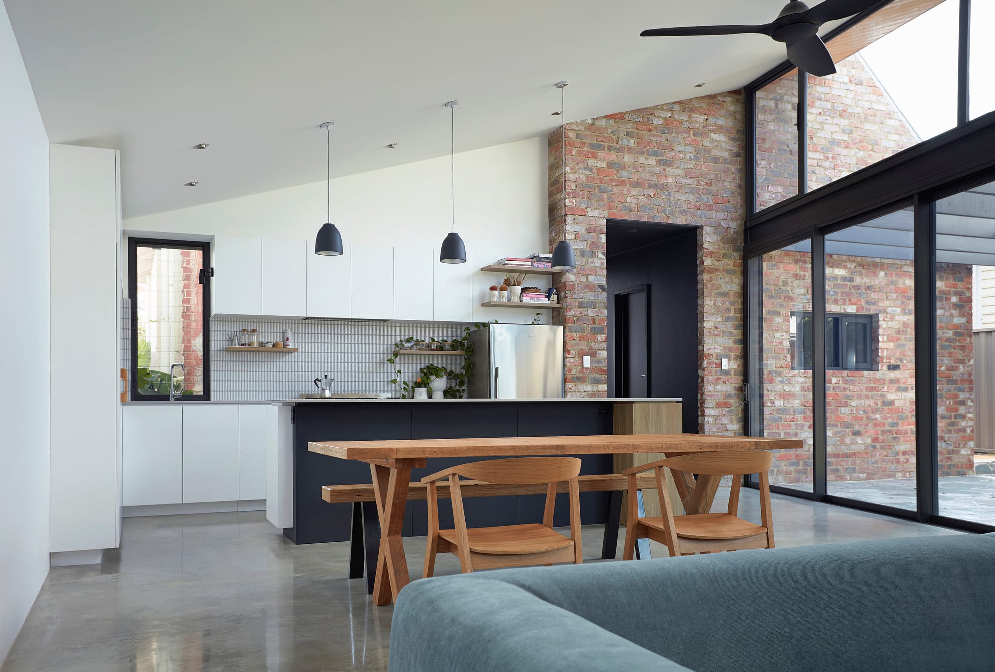 Displayed is a stylish kitchen with modern amenities, white cabinetry, and a large wooden dining table complemented by matching chairs. The design incorporates exposed brick walls, blending industrial and contemporary elements. Large, black-framed windows and doors open to the exterior, offering views of the adjacent outdoor space and providing a bright and airy atmosphere.