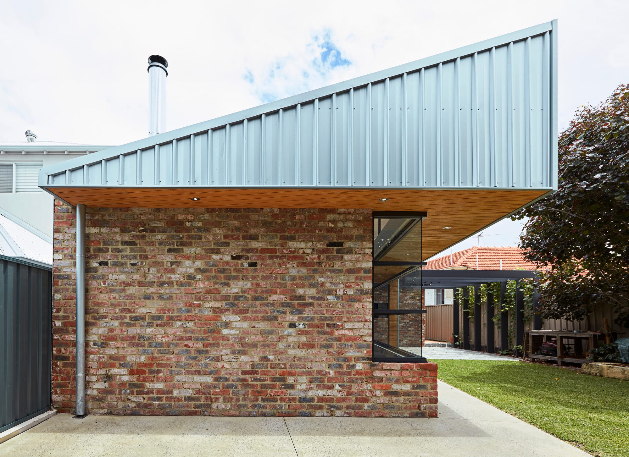 Sharp House by Steelehouse Architecture.The final image presents the rear of a contemporary house, characterized by its corrugated metal roof and warm wooden soffits. The design is a harmonious combination of industrial and natural elements, featuring large glass windows for an indoor-outdoor living experience. The backyard is tidy, with a narrow garden bed lining the brick base of the home.