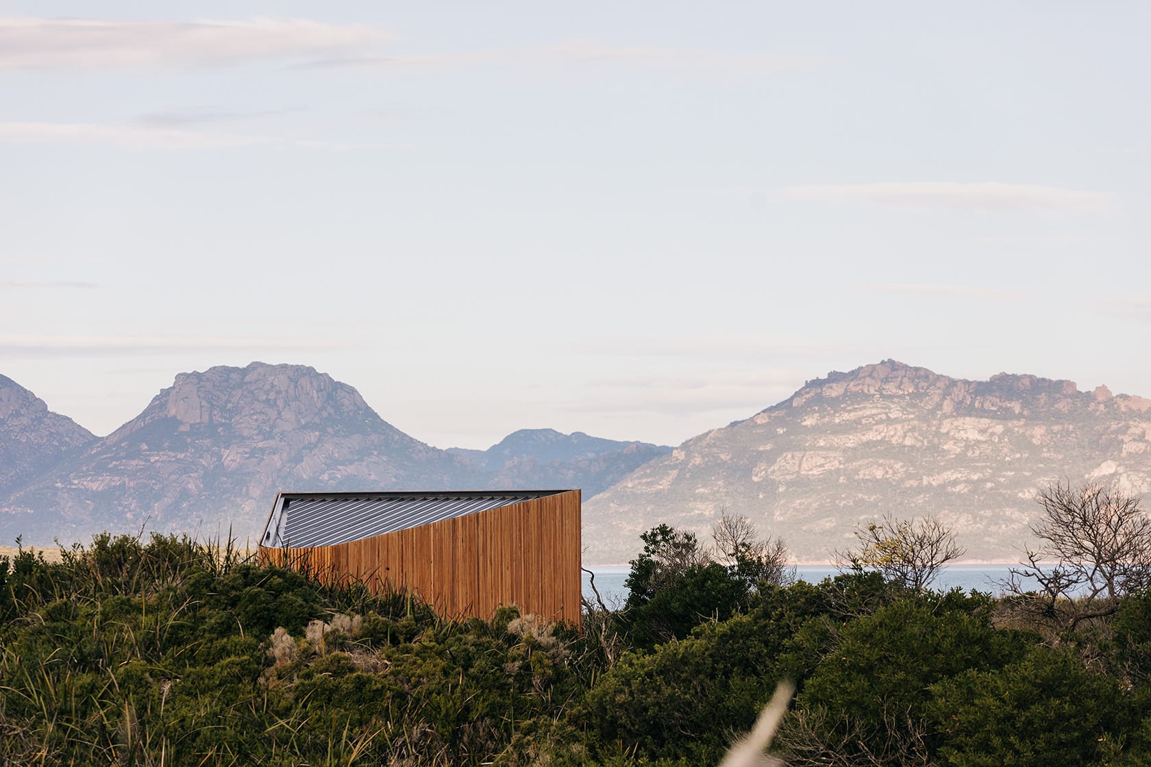 Studio Tasmania in Dolphin Sands, Tasmania. A sleek, architecturally modern structure with vertical wooden slats stands in contrast to the wild, undulating dunes of Dolphin Sands. The building's unique angular form juxtaposes the organic shapes of the distant rugged mountains, under a vast sky with soft cloud patterns.