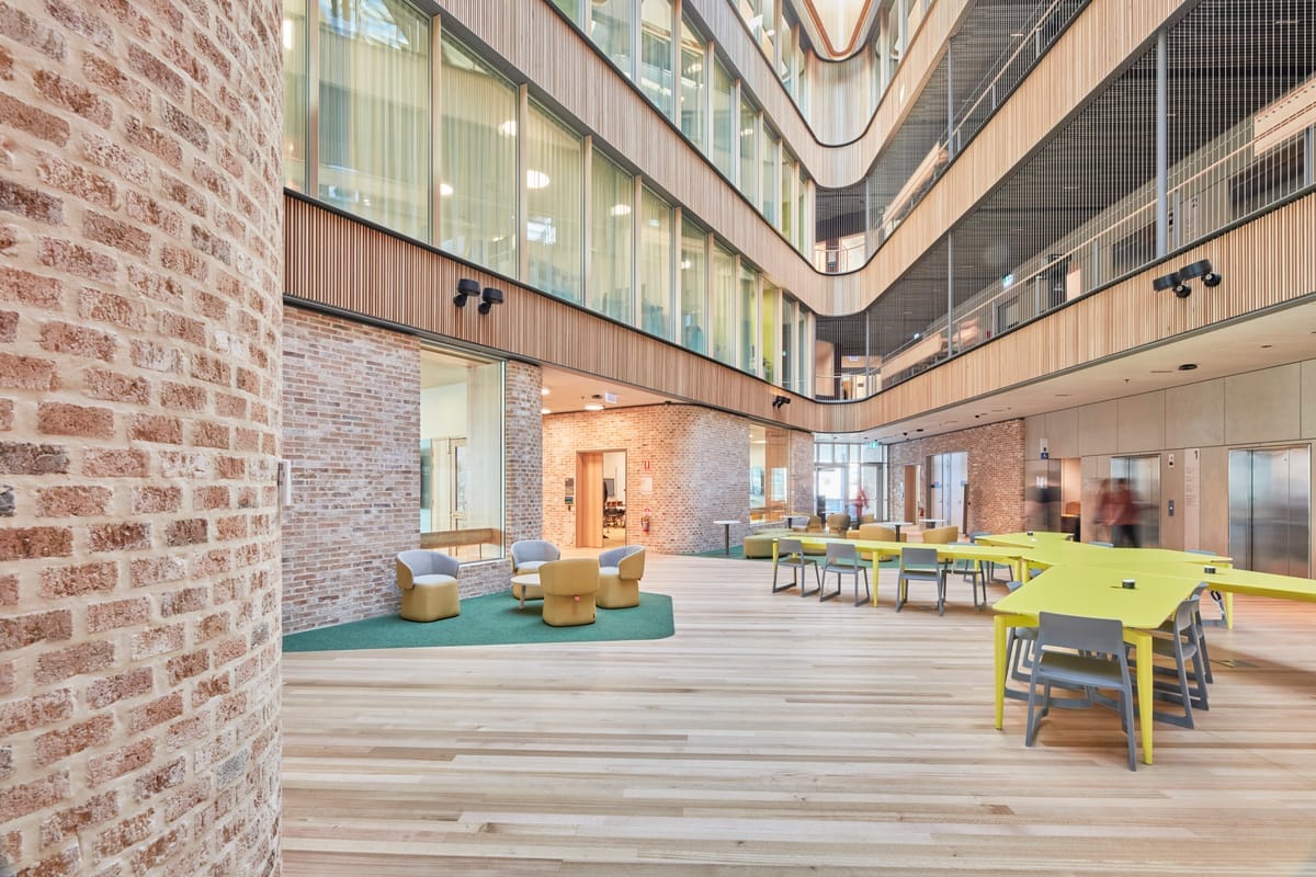 Tasmanian Timber: River's Edge. Photography: Dave Groves.A spacious interior corridor flanked by plush, mustard-colored seating and round, gray tables on one side and a wood-paneled wall on the other. The ceiling features unique, abstract-shaped light fixtures, while the far end showcases a windowed facade and mezzanine levels, adding depth to the modern architectural space.